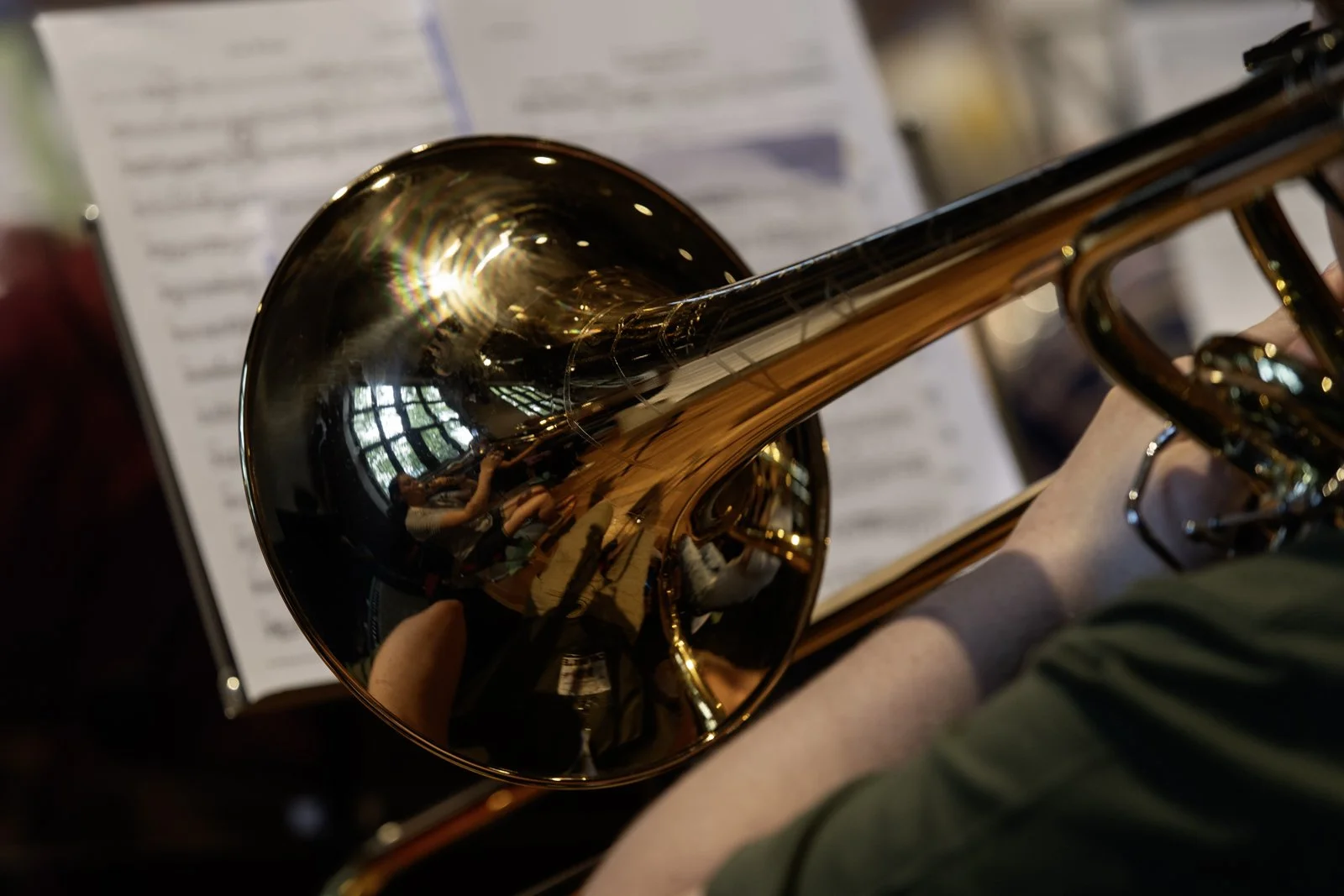 A close-up of a person playing a brass trumpet, with sheet music in the background and a reflection of the room in the instrument's bell, showing multiple people and windows.
