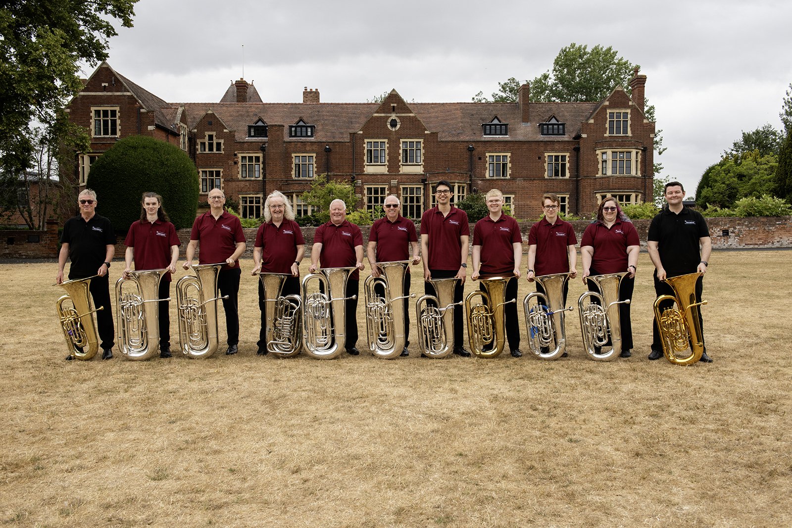 A group of ten people standing outdoors on a grassy field, holding musical instruments, in front of a large brick building with trees around.