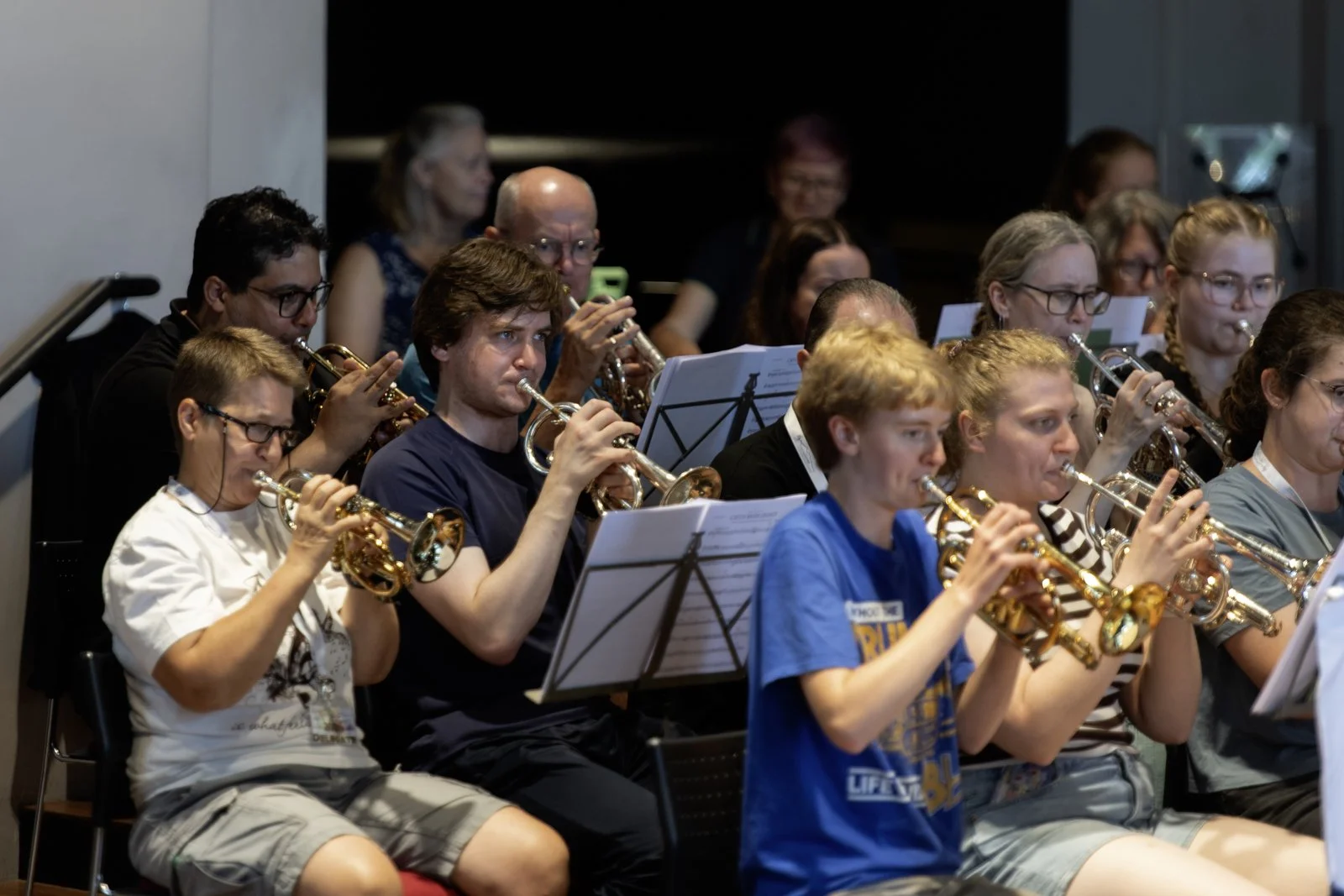 Group of musicians playing saxophones, seated in a music rehearsal or performance setting, with sheet music on stands in front of them.