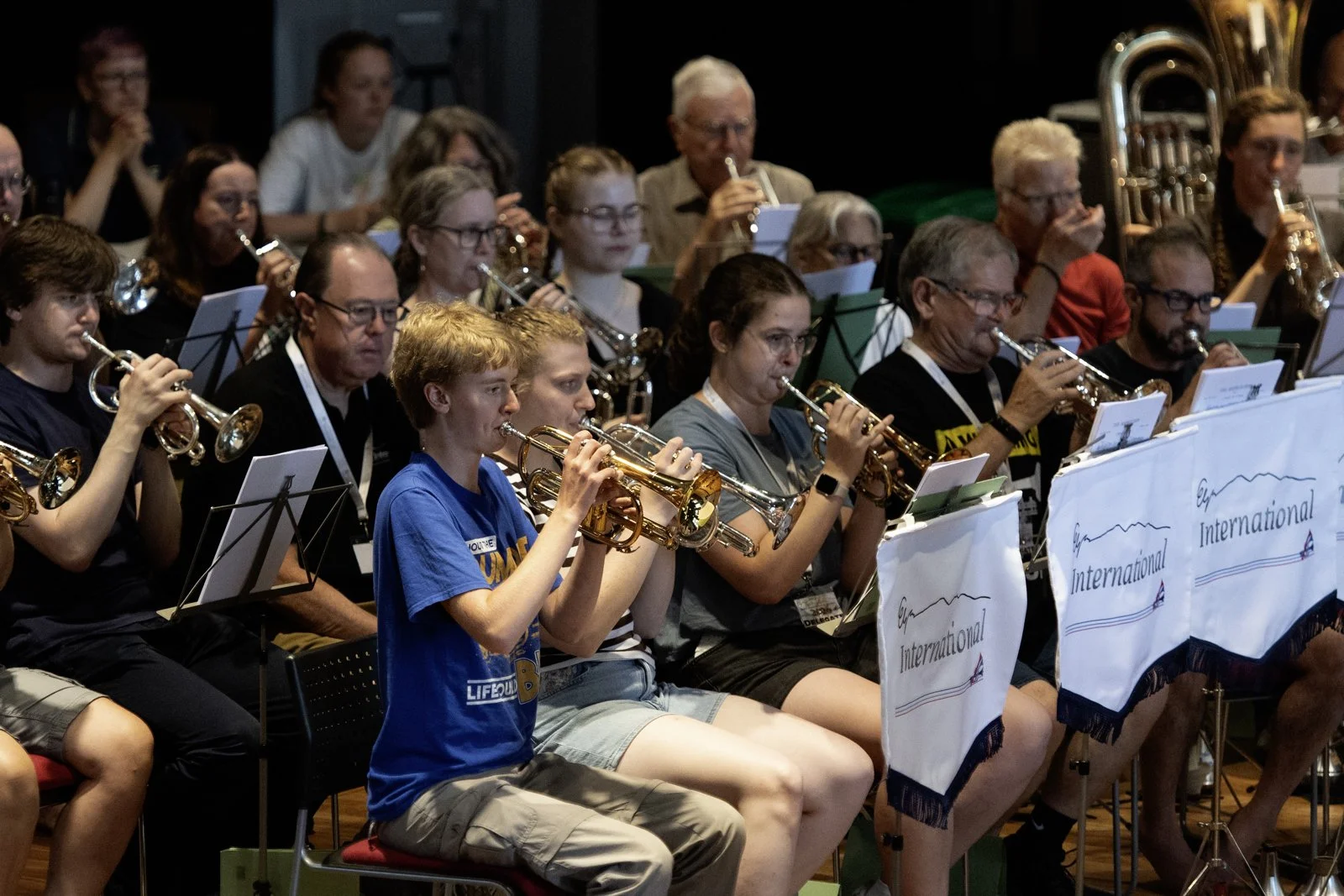 A group of people, mostly older adults and children, playing trumpets during an orchestral rehearsal or performance, with some reading music sheets and wearing music stands with banners that read 'Alaska International.'
