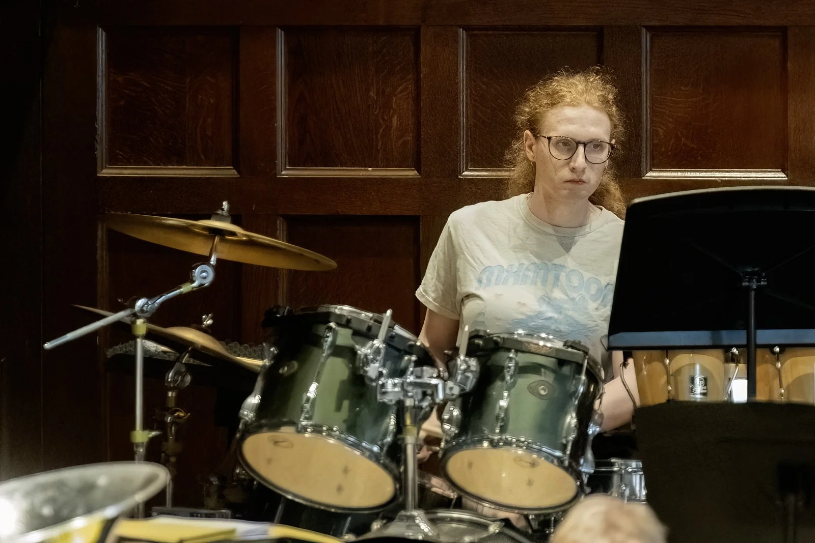 A person with glasses and curly, reddish hair playing a drum set, with a dark wooden panel background.
