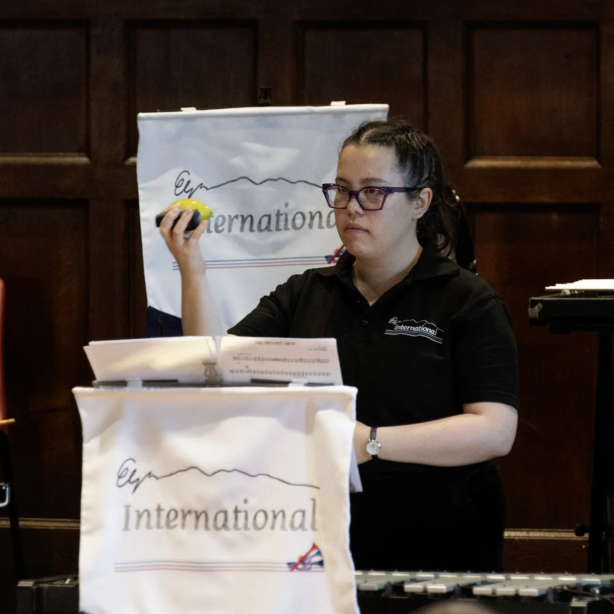 A woman with glasses and dark hair tied back, wearing a black shirt and a wristwatch, is holding a lemon in front of her face at an event. She is standing in front of a music stand with sheet music, and documentation. In the background, there is a ba