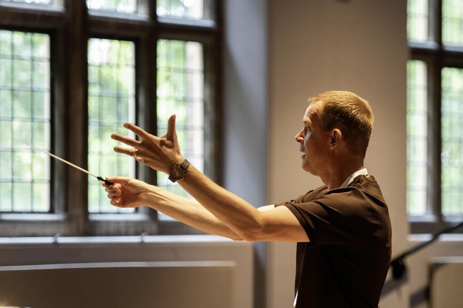 A man conducting an orchestra or choir, gesturing with both hands against a background of large windows.