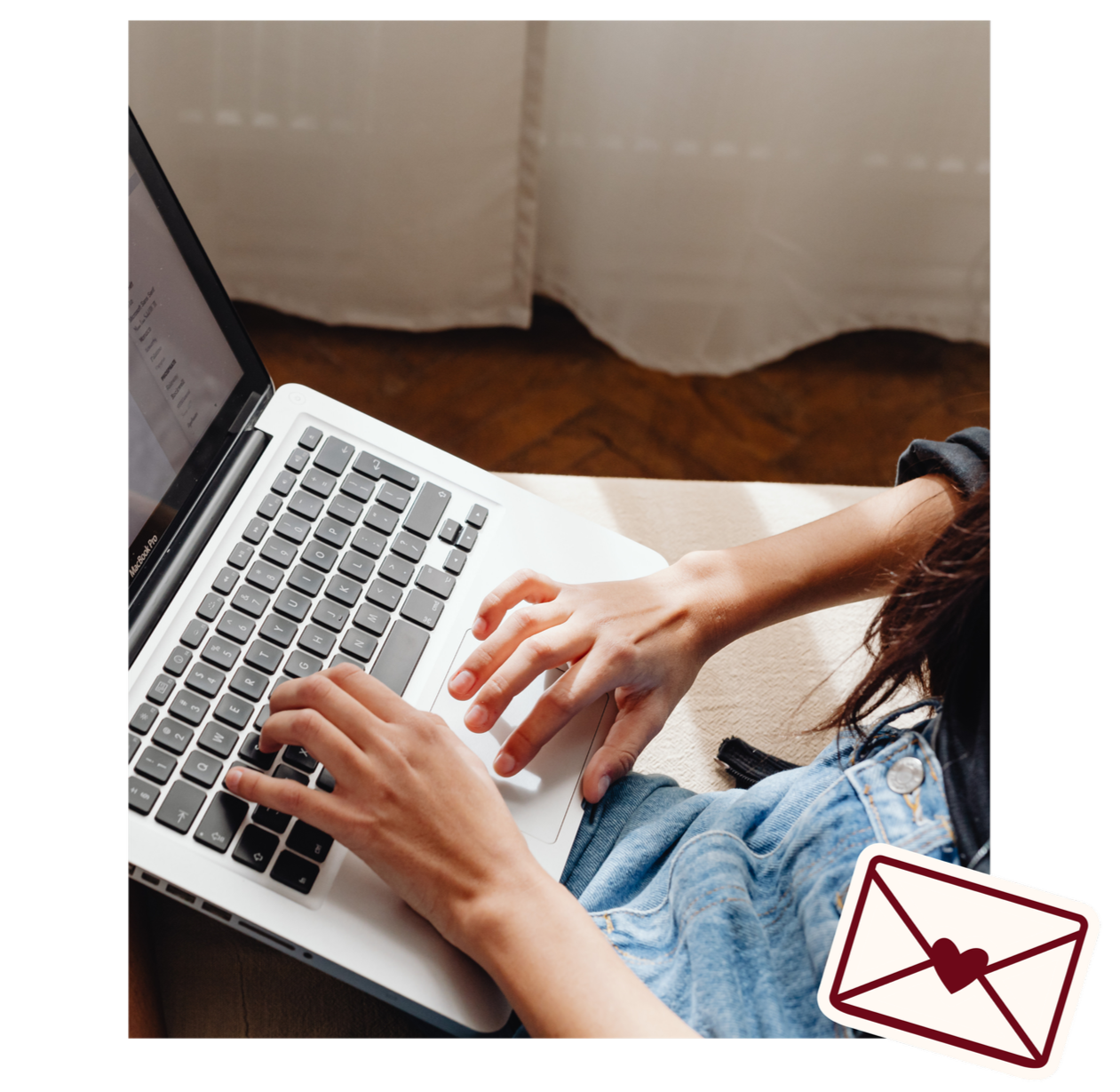 A person typing on a laptop keyboard. The person appears to be wearing a denim jacket and black long sleeves, with part of their brown hair visible. The laptop is on a beige surface, and a curtain and wooden floor are visible in the background. There is a sticker with an envelope and heart in the bottom right corner of the image.