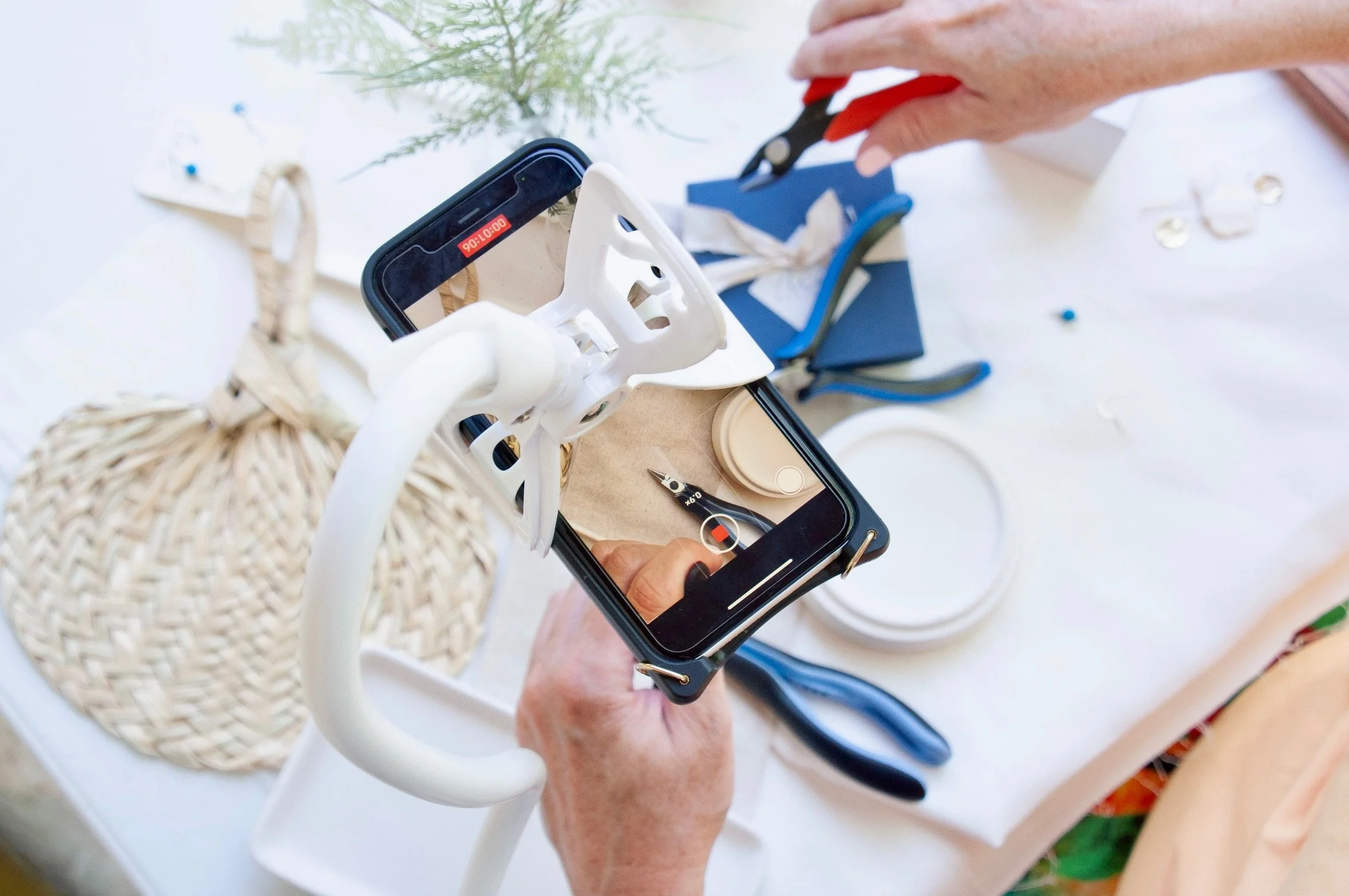 Person using a smartphone to take a photo of a wicker basket with a white lab coat, scissors, and other craft supplies on a table.