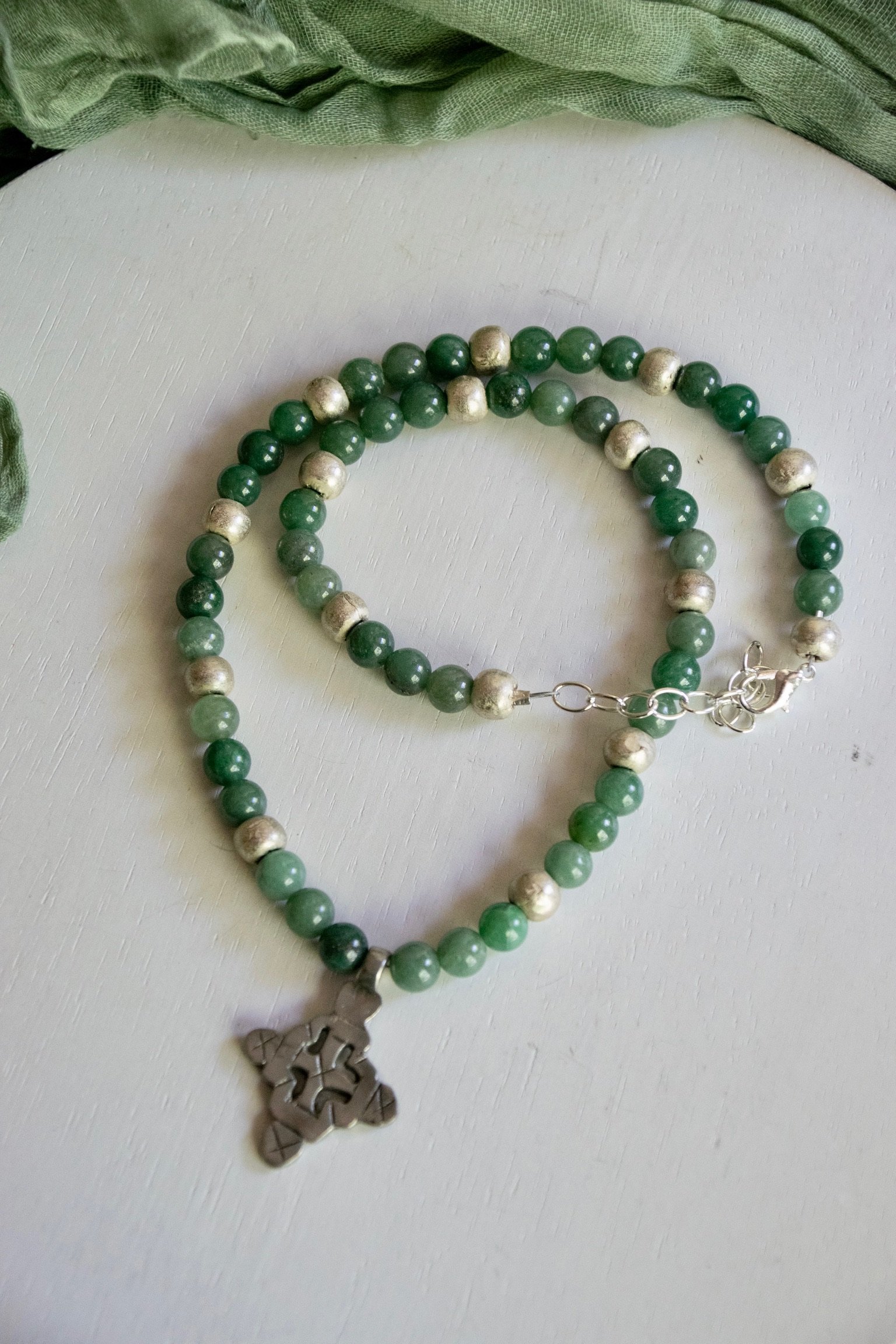 Green beaded necklace with silver accents and a silver pendant shaped like a snowflake, lying on a white surface with a piece of green fabric partially visible above.