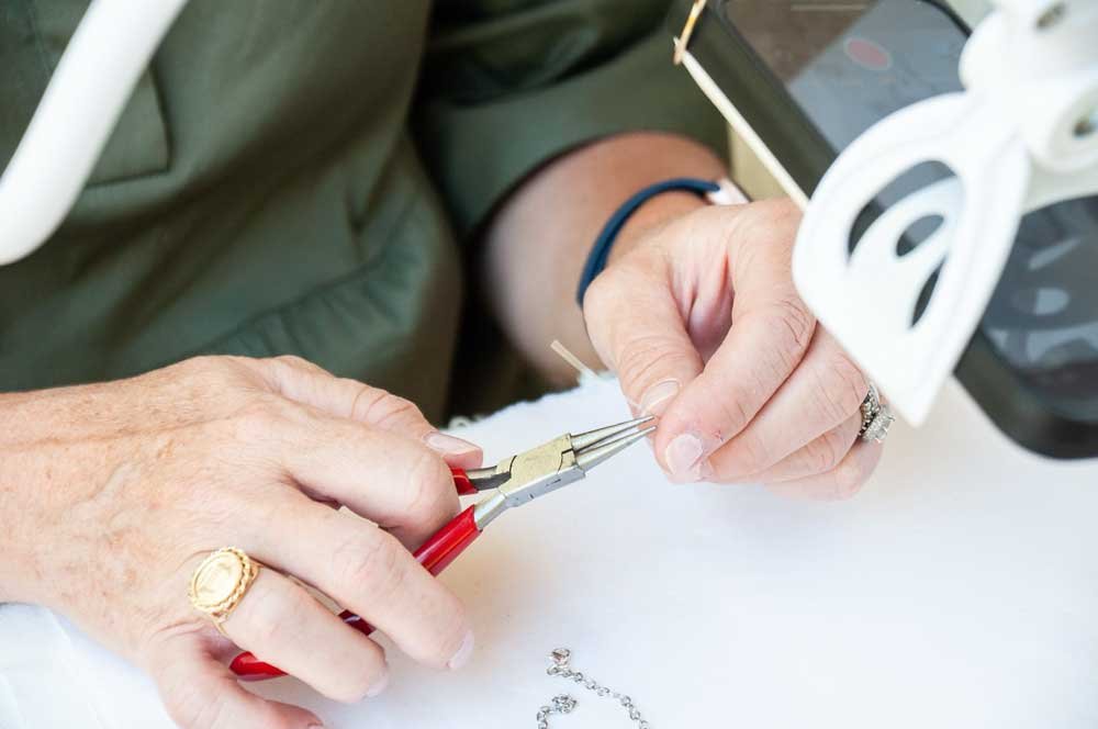 Hands of a person working with fine jewelry, using tweezers to hold a small wire or component, with jewelry pliers in hand, on a white surface.