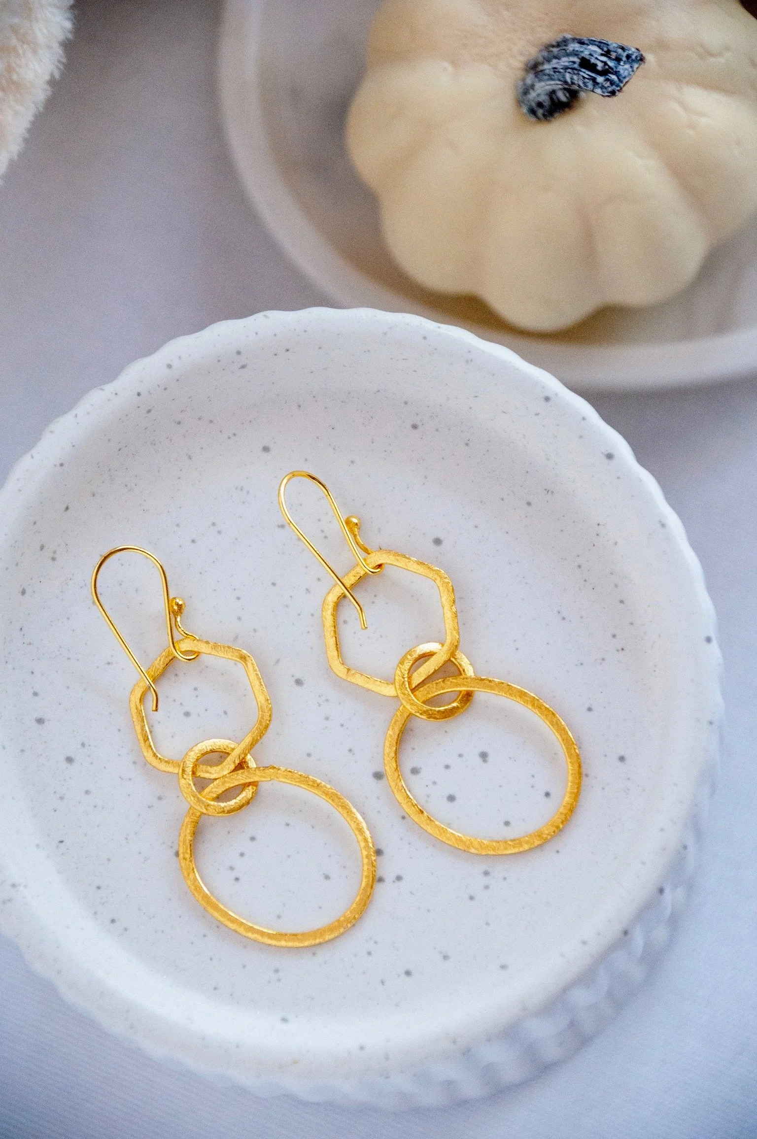 Gold geometric-shaped earrings on a white speckled dish, with a blurred background of a soft beige textile and a dish with an ice cream scoop with a black and white topping.