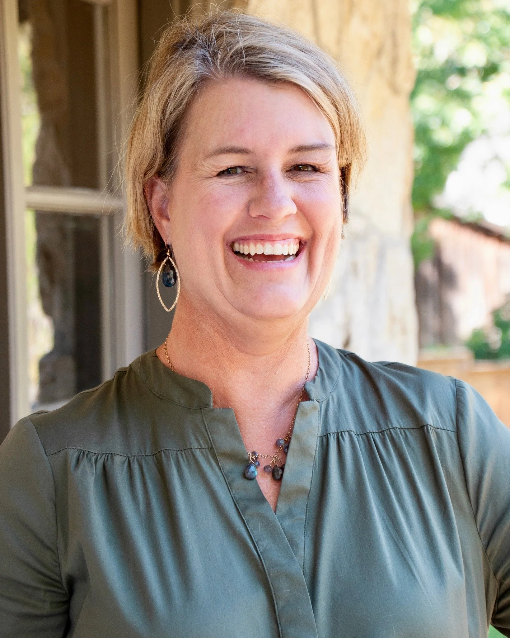 A smiling woman with short blonde hair, wearing a dark green top, earrings, and a beaded necklace, standing outdoors beside a stone wall and window, with trees in the background.