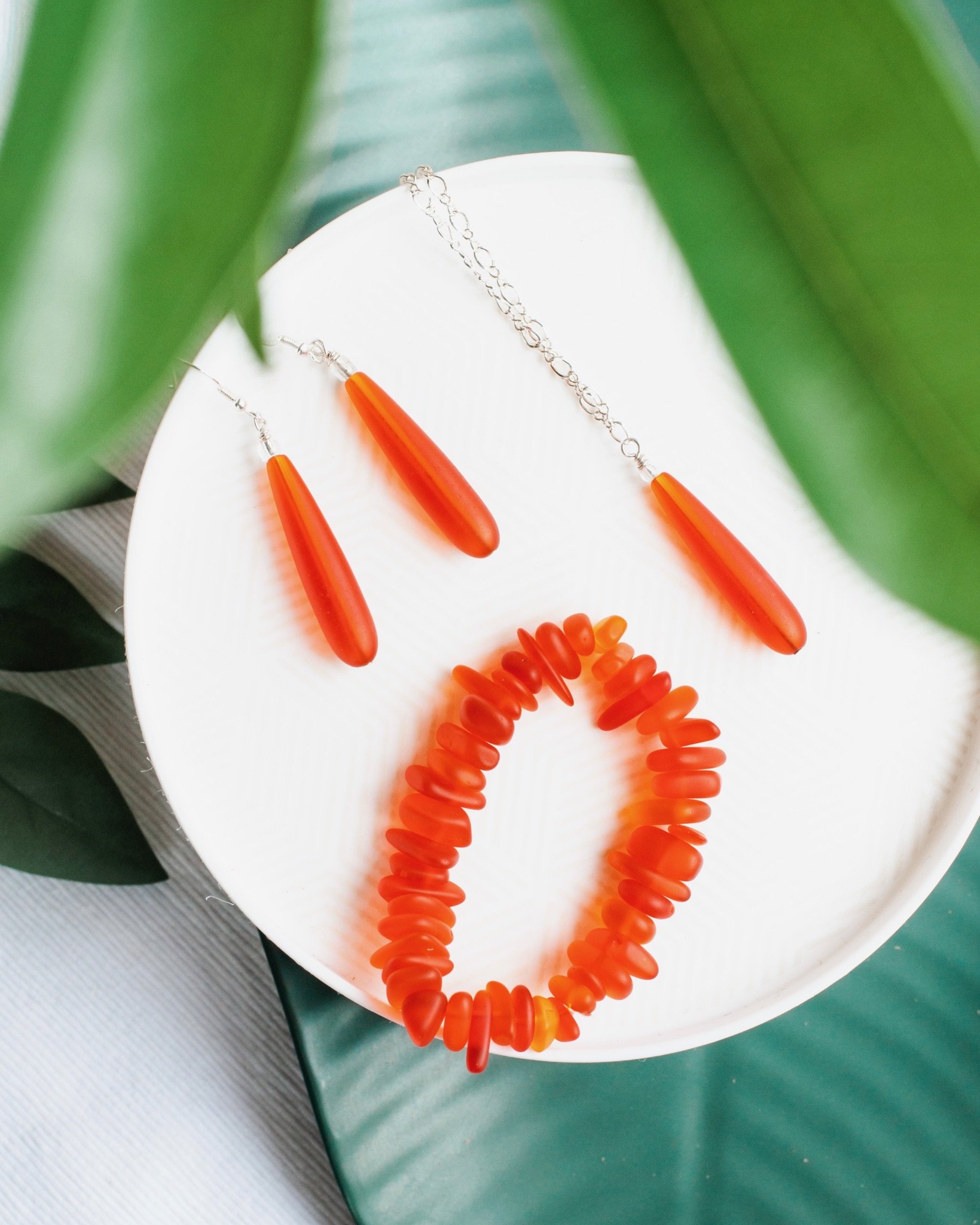 Jewelry with orange beads and chain on a white surface with green leaves in the foreground.