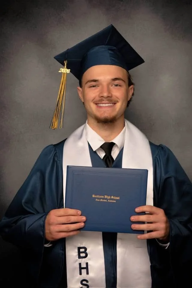 A young man dressed in a blue graduation gown and cap with a gold tassel, holding a diploma in front of him, smiling at the camera.