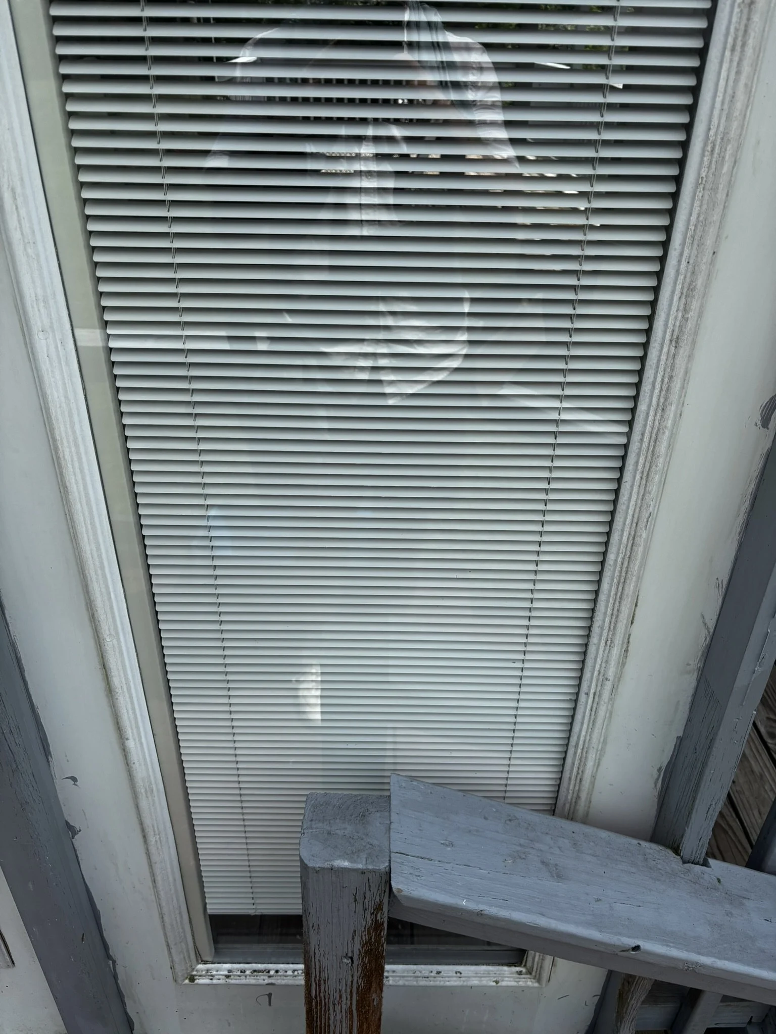 A vent with horizontal slats on a door or window, with a white frame. The vent cover is white, and above it, part of the window with closed blinds is visible. There is some weathered wood trim and a wooden post at the bottom of the image.