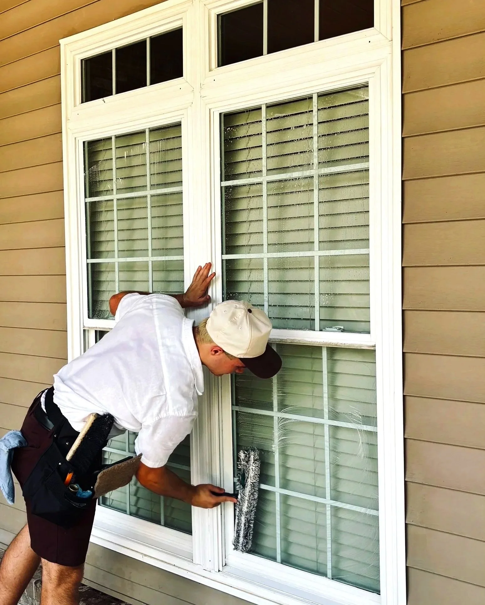 Two men are cleaning a large window with white frames on a house with beige siding. One man is wearing a white shirt and a baseball cap, and the other is wearing a white shirt and a tool belt. The window panes are divided into smaller sections with horizontal and vertical bars.