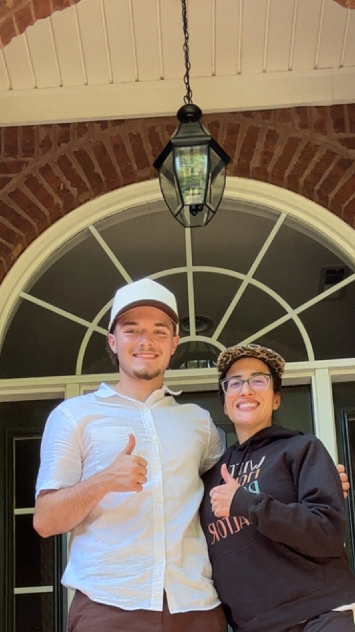 Two young men smiling and giving thumbs up in front of a large window and brick wall, with a hanging lantern above.
