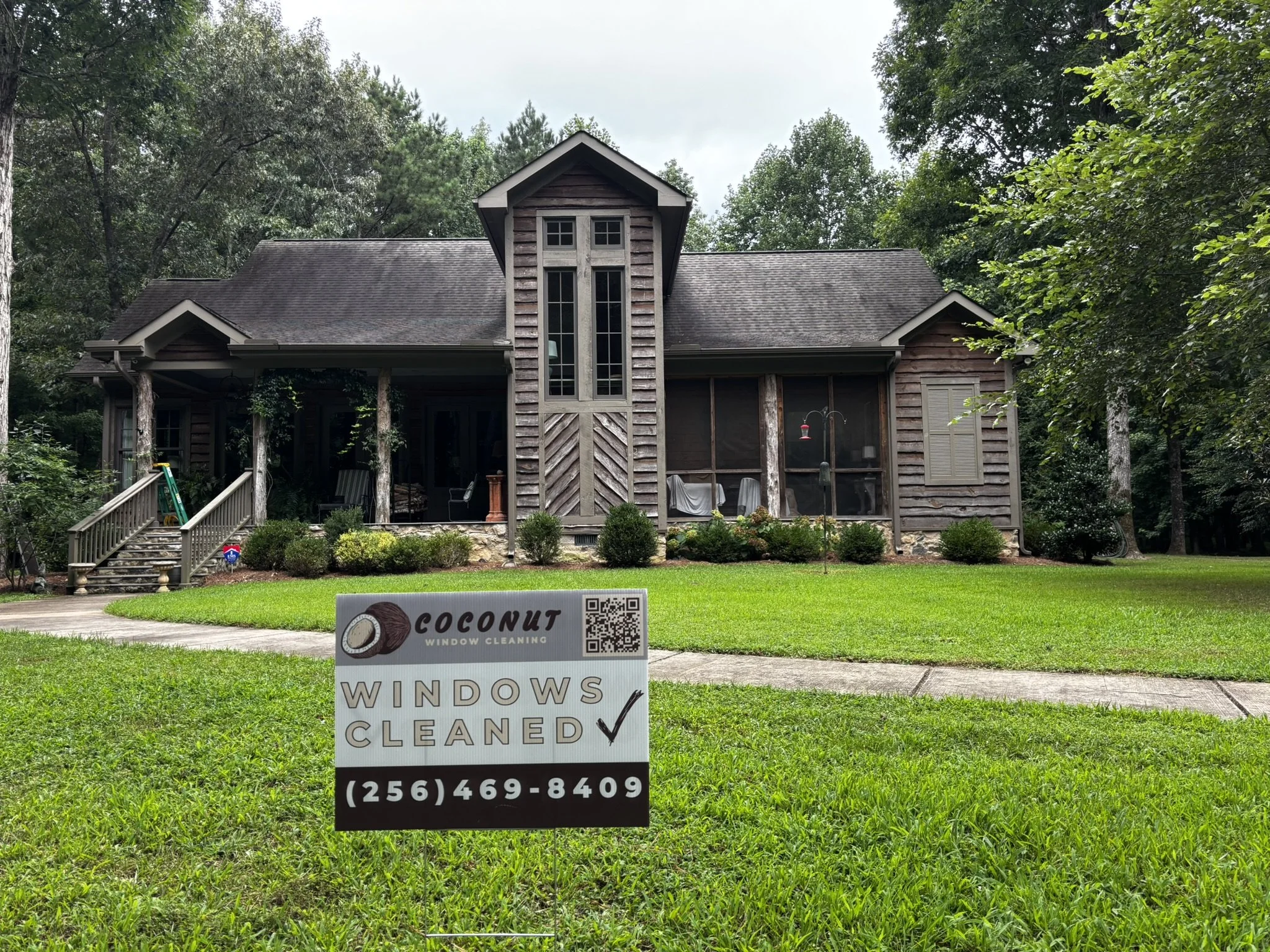 A house with a wooden exterior surrounded by trees and a well-maintained lawn. A sign in the front yard advertises window cleaning services, displaying the word 'Coconut' with a picture of a coconut, a checkmark, and the phone number (256) 469-8409.