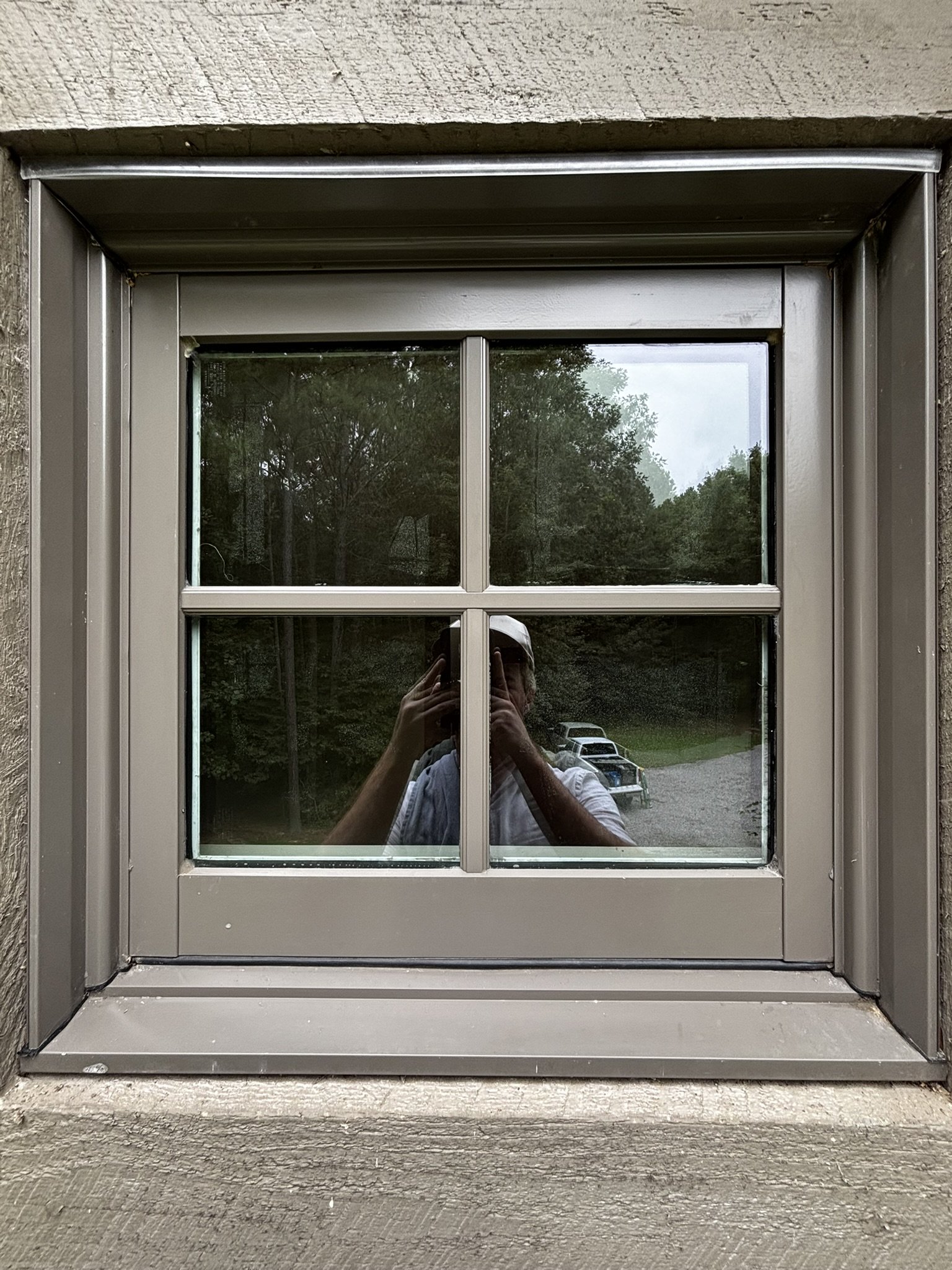 Close-up of a house window with white frame and black lattice, reflecting trees and a person taking a photo, with a wooden windowsill and brick wall surrounding.