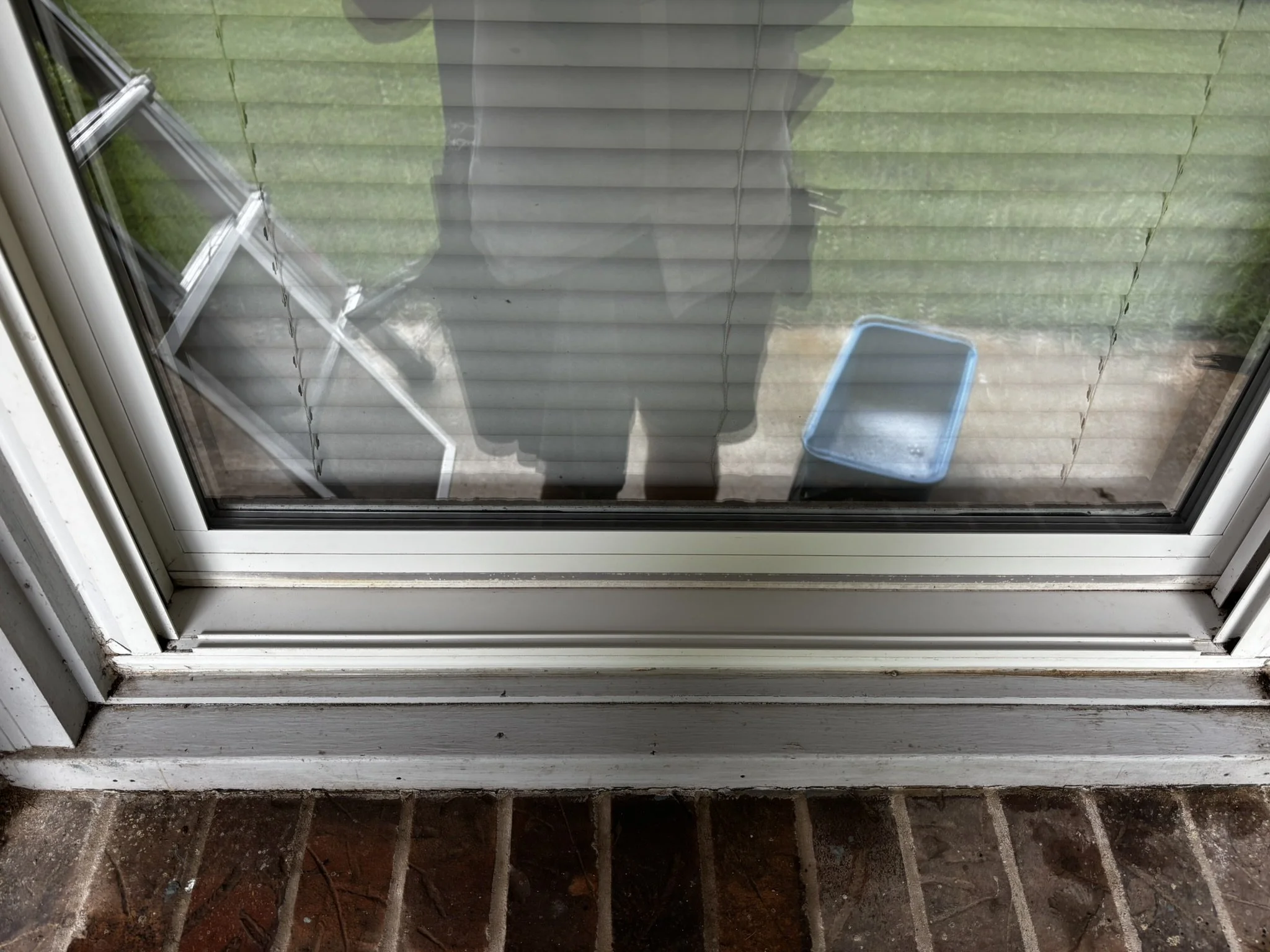 View of a glass door with white framing, showing a patio with brown tiles and a grill inside. Outside, there is a small green lawn and a plastic tray on the ground.