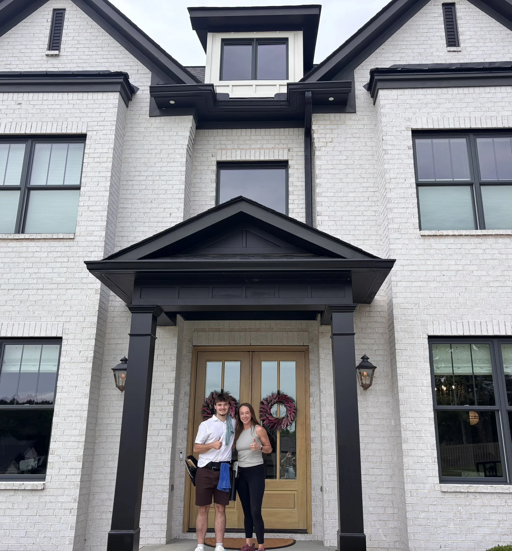 A young man and woman standing in front of a modern white brick house, smiling and giving thumbs up. The house has large windows, a wooden front door with glass panels, black accents, and two wall lanterns on either side of the door. The porch is covered with a black roof supported by black pillars, and a decorative pink wreath hangs on the door.