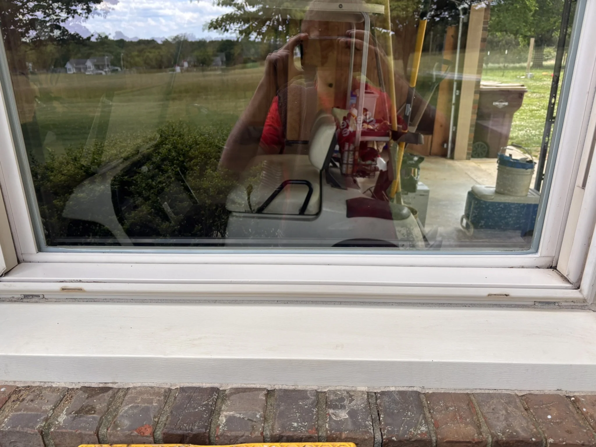 Outdoor scene viewed through a window, showing a grassy yard with trees, houses, and mountains in the background. The reflection of a person taking a photo with a smartphone can be seen on the glass.