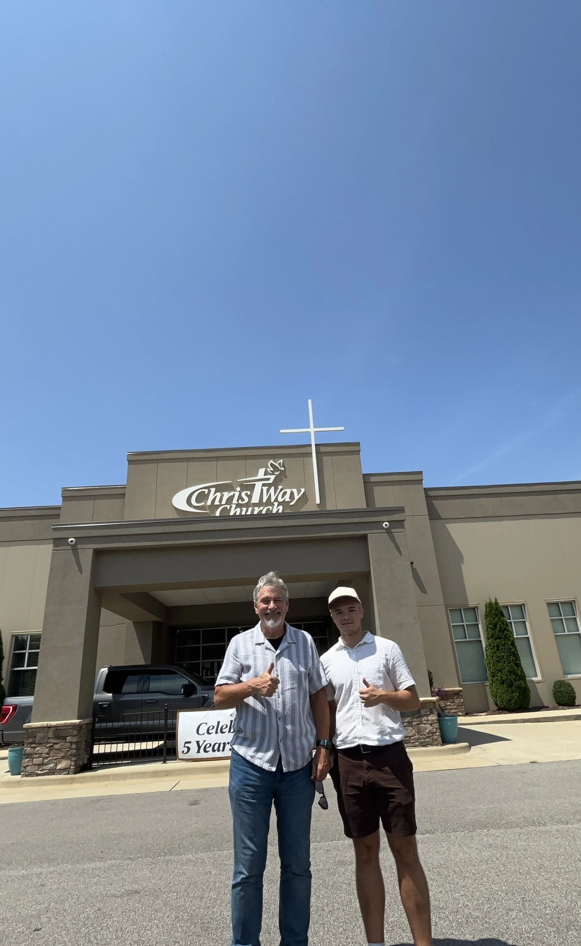 Two men standing in front of ChristWay Church, celebrating 5 years, with a blue sky overhead and a cross on the church building.