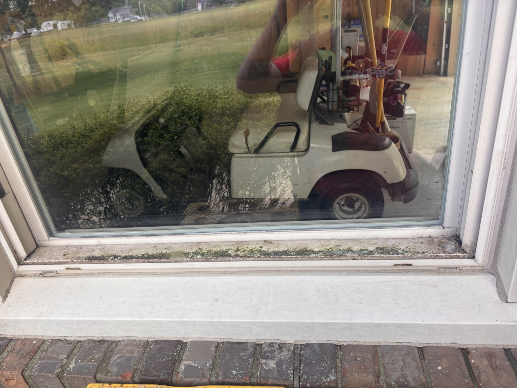 View through a window showing a golf cart parked inside a garage with tools and equipment, reflecting a grassy outdoor area with trees and neighboring houses.