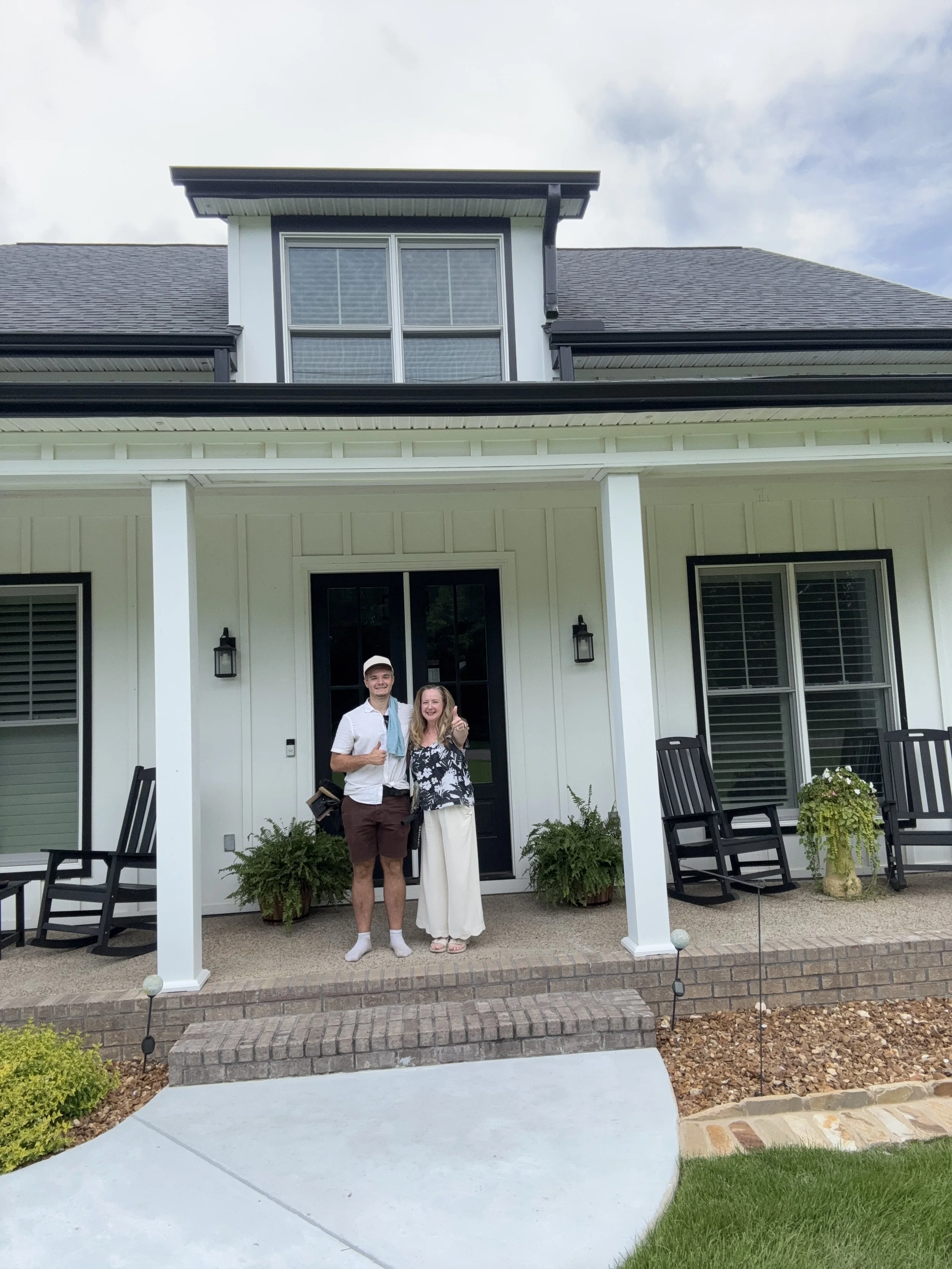 A man and a woman standing in front of a white house with black accents, smiling and giving thumbs up. The house has a black front door, large windows with white blinds, black chairs outside, and potted plants on either side of the entrance. The patio area has a curved concrete walkway, a brick-bordered flower bed, and decorative garden lights.