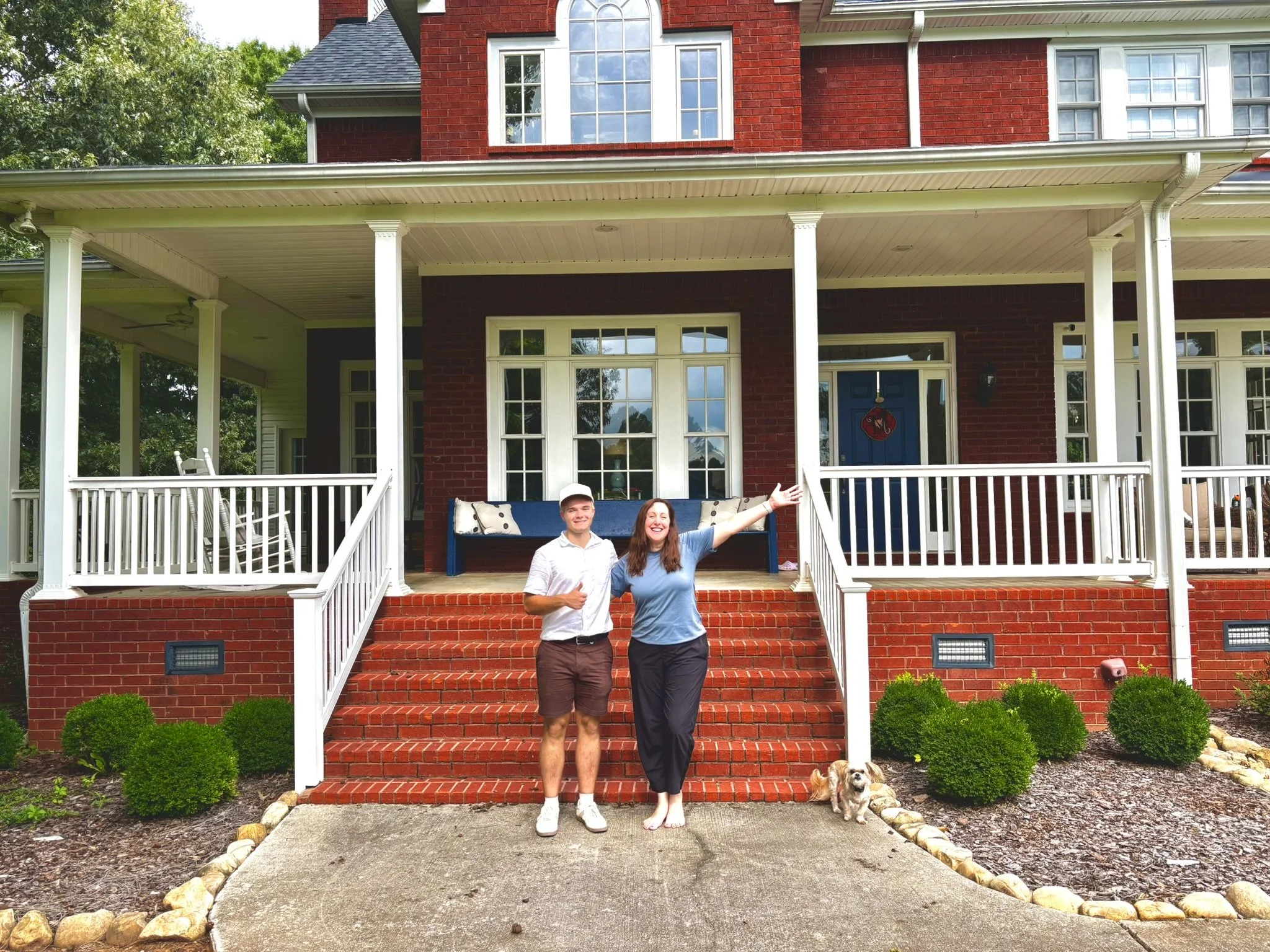 A smiling man and woman stand on the front steps of a red brick house with a large porch. The woman is pointing outward and holding a small dog at her feet. The man has his thumb up. The house has white trim, multiple windows, and a decorated door, with greenery and bushes in the yard.