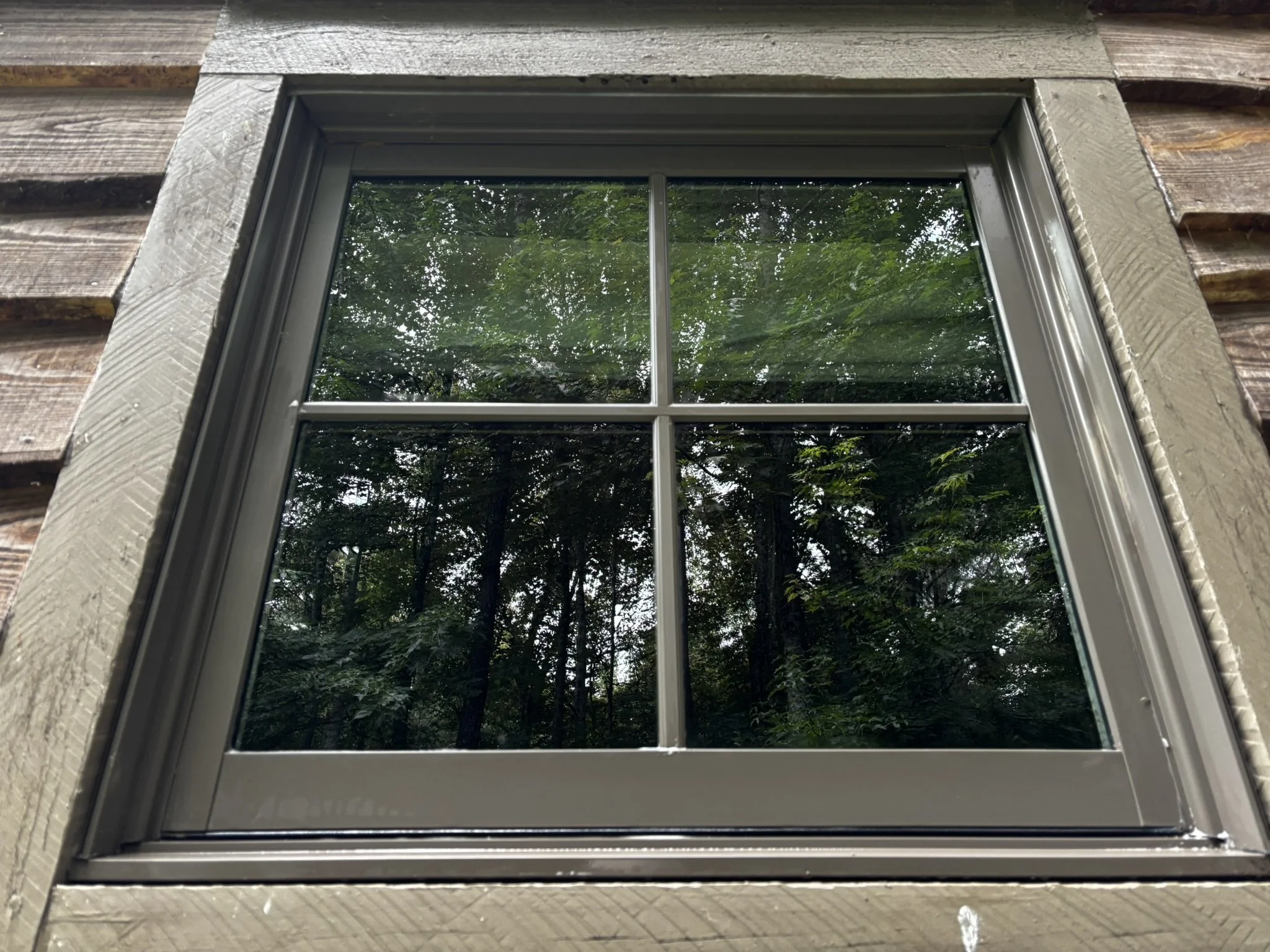 Close-up view of a window with a grid of four panes, reflecting green trees and foliage outside, surrounded by wooden and stone building materials.