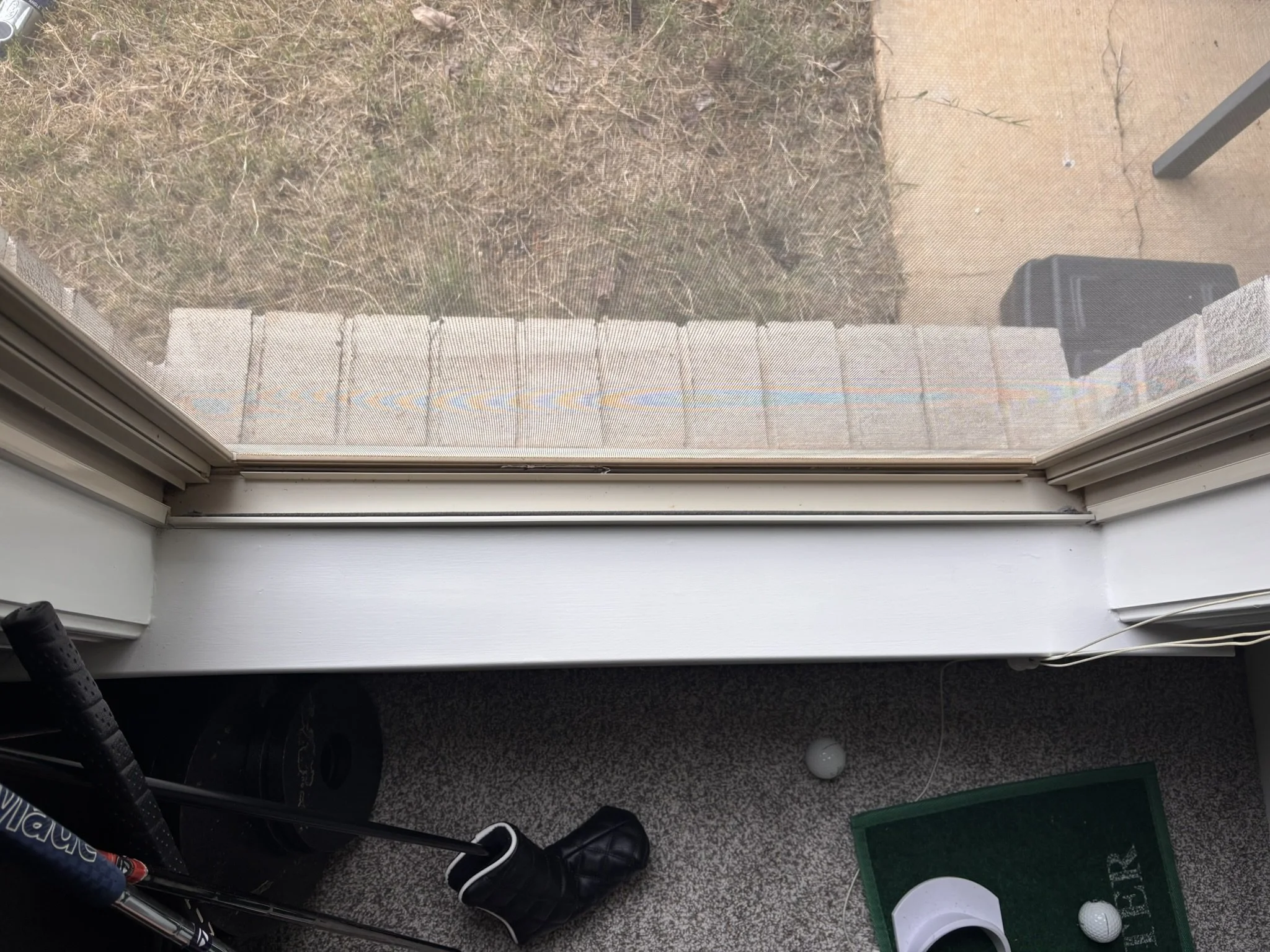 View through a window screen looking out onto a patio with a dry grassy yard, a small pizza box, and golf balls on the ground. A golf club and a boot are partially visible in the foreground.