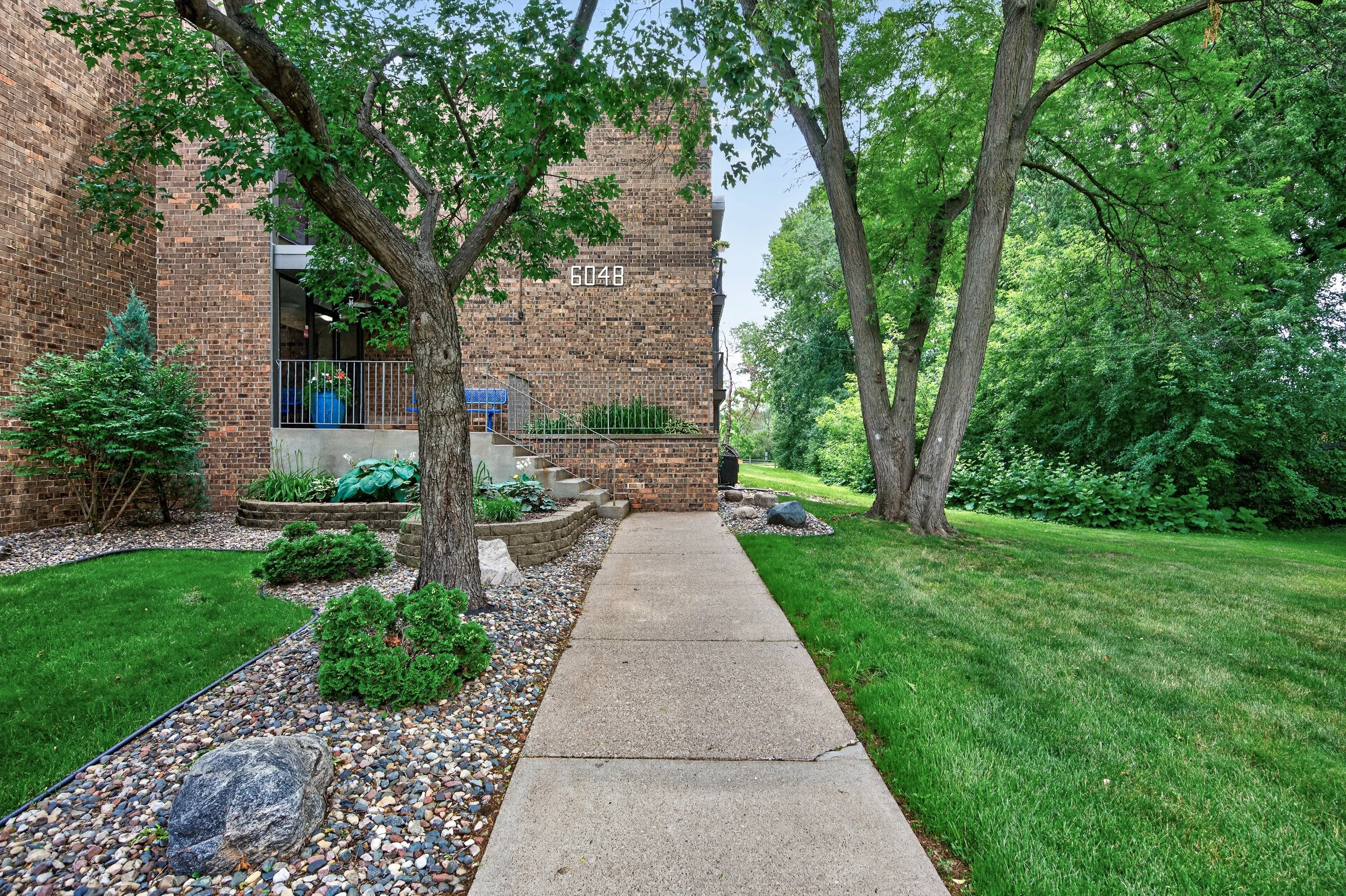 A sidewalk leading to a residential building at Eastgate Apartments in White Bear Lake, MN, surrounded by green grass, trees, and landscaped garden beds with rocks and plants.