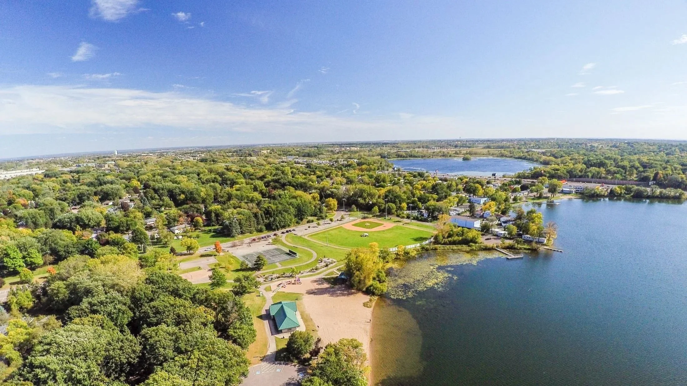 An aerial view of a large park from Eastgate Apartments in White Bear Lake, MN with a baseball field, a playground, and water bodies surrounded by dense green trees on a sunny day.
