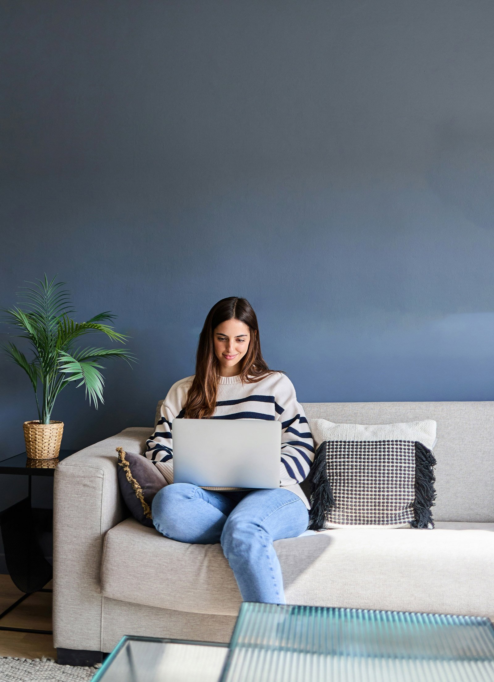 Woman with long brown hair sitting on a beige couch, using a silver laptop, with a gray and black pillow to her right. She wears a white sweater with navy blue stripes and light blue jeans. Behind her is a dark blue wall, with a green potted plant on her left and a black side table.