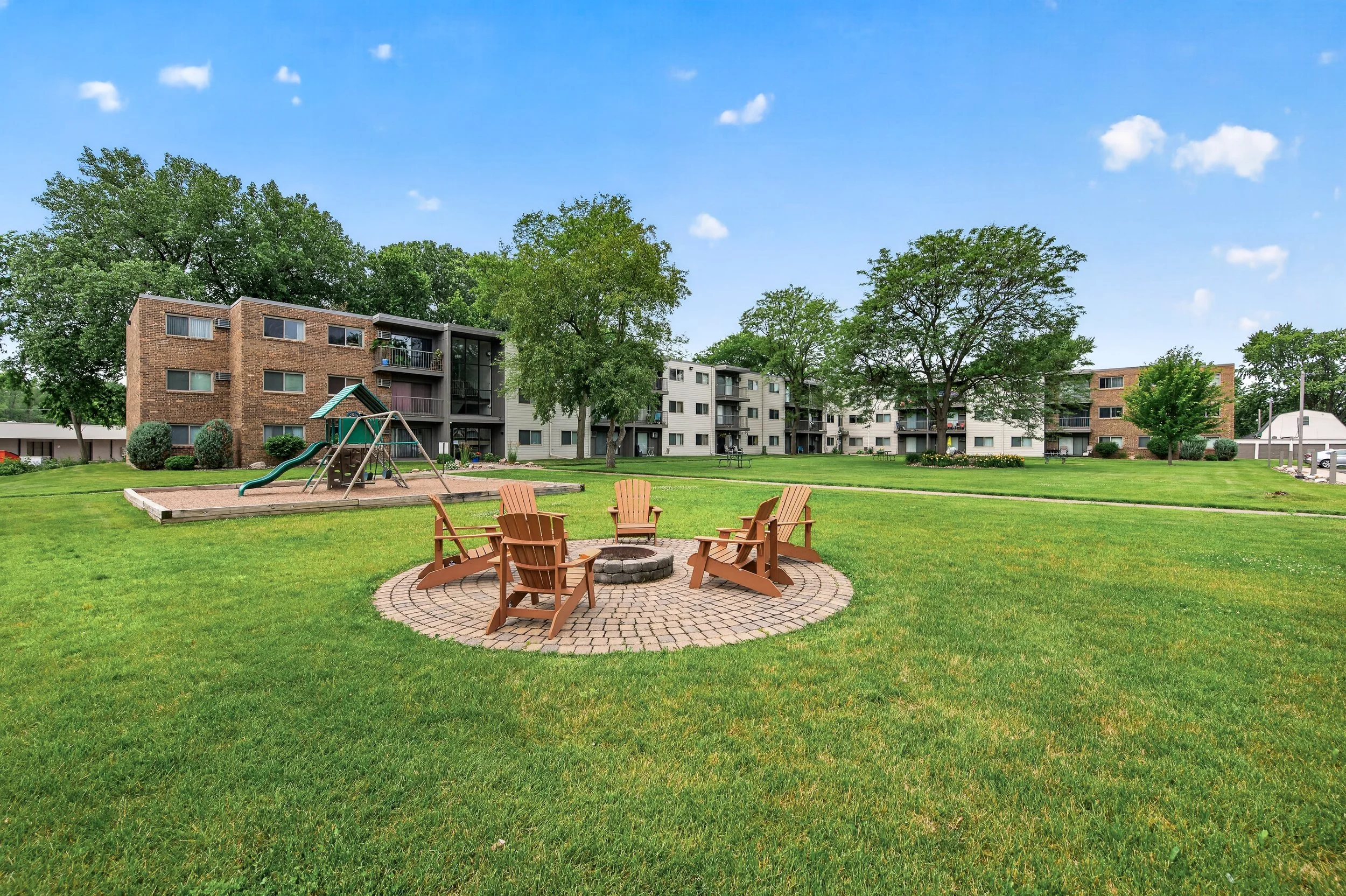Eastgate Apartment complex with a playground, wooden chairs surrounding a fire pit, large trees, and well-maintained lawn under a partly cloudy sky.