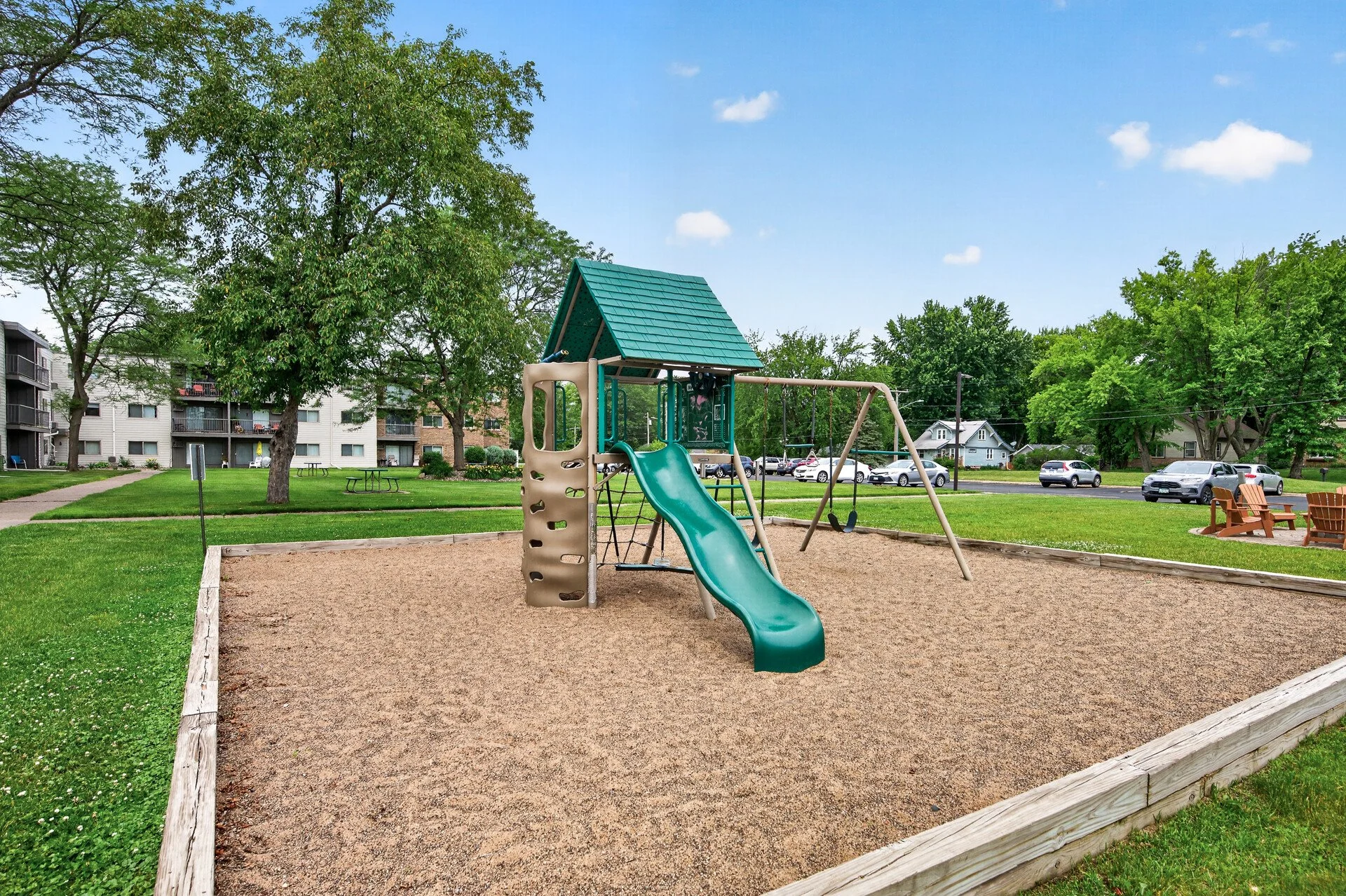 Children's playground with slide and swings in a park surrounded by green grass and trees, with Eastgate Apartment buildings and parked cars in the background on a sunny day.
