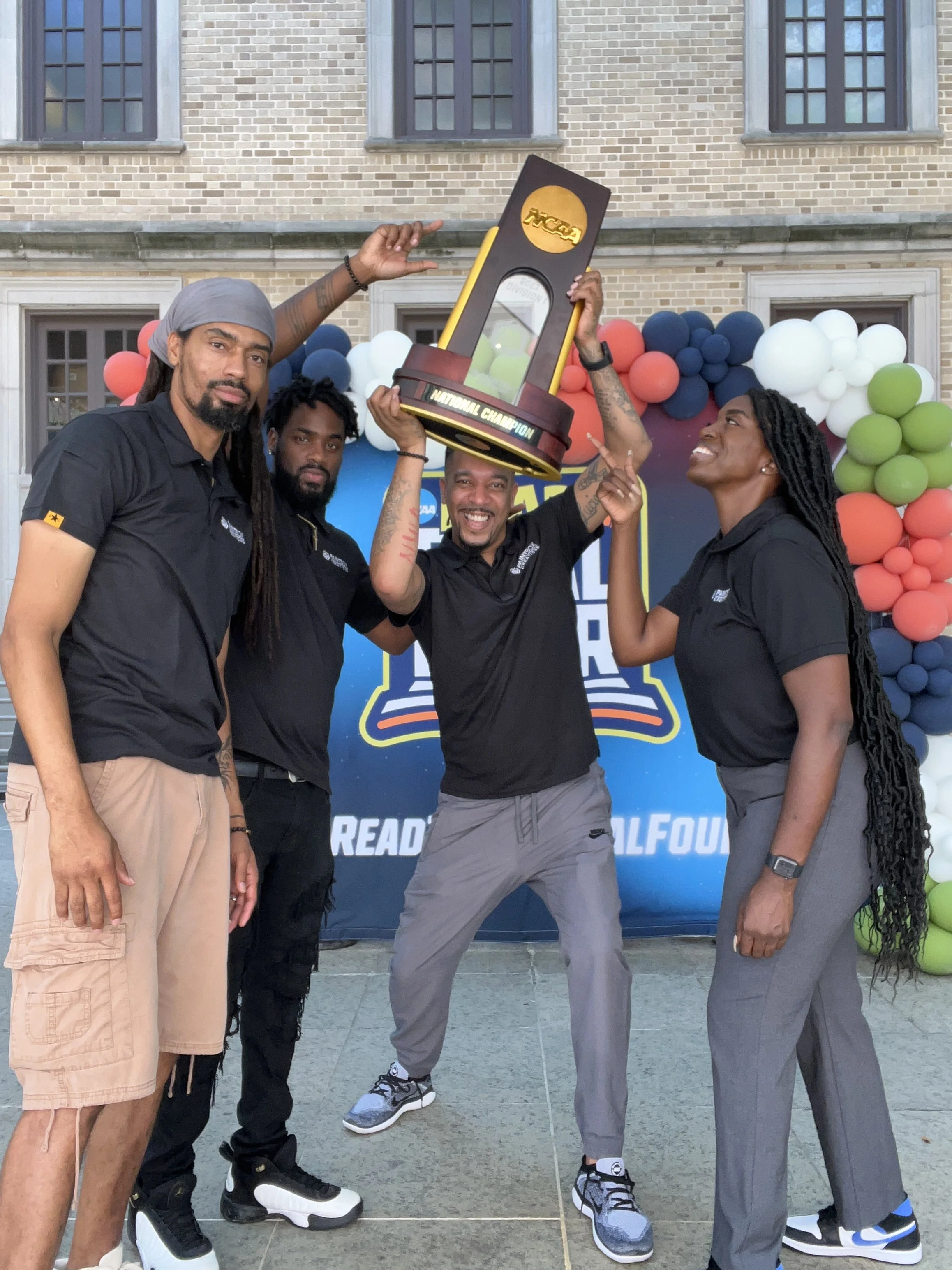 Four men standing in front of a brick building with balloons and a banner, celebrating a sports championship. One man is holding a large trophy-shaped award labeled 'NCAA' and 'National Champion'.