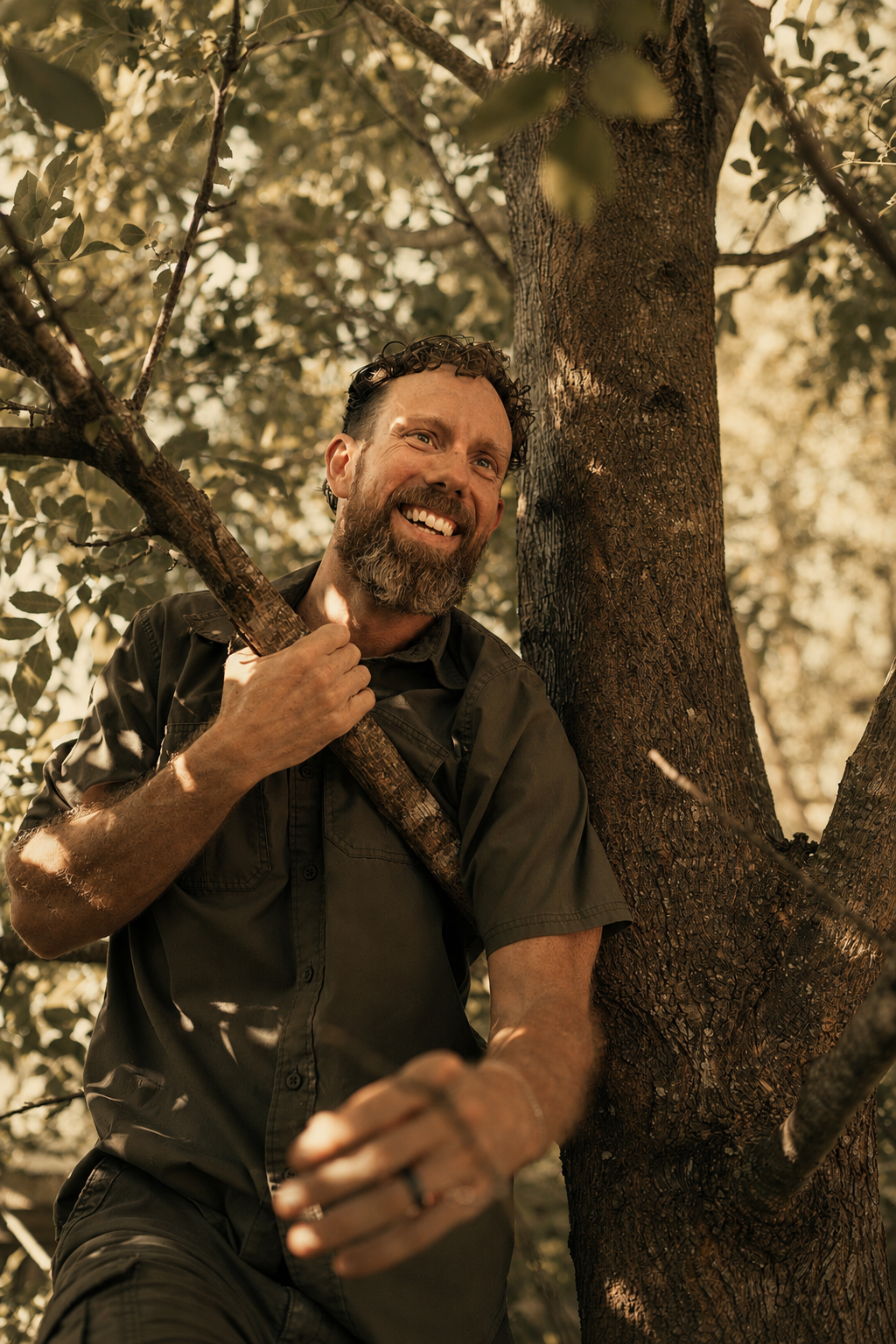 A man with a beard and curly hair smiling while standing next to a tree, holding a branch with one hand, in a forest or wooded area.