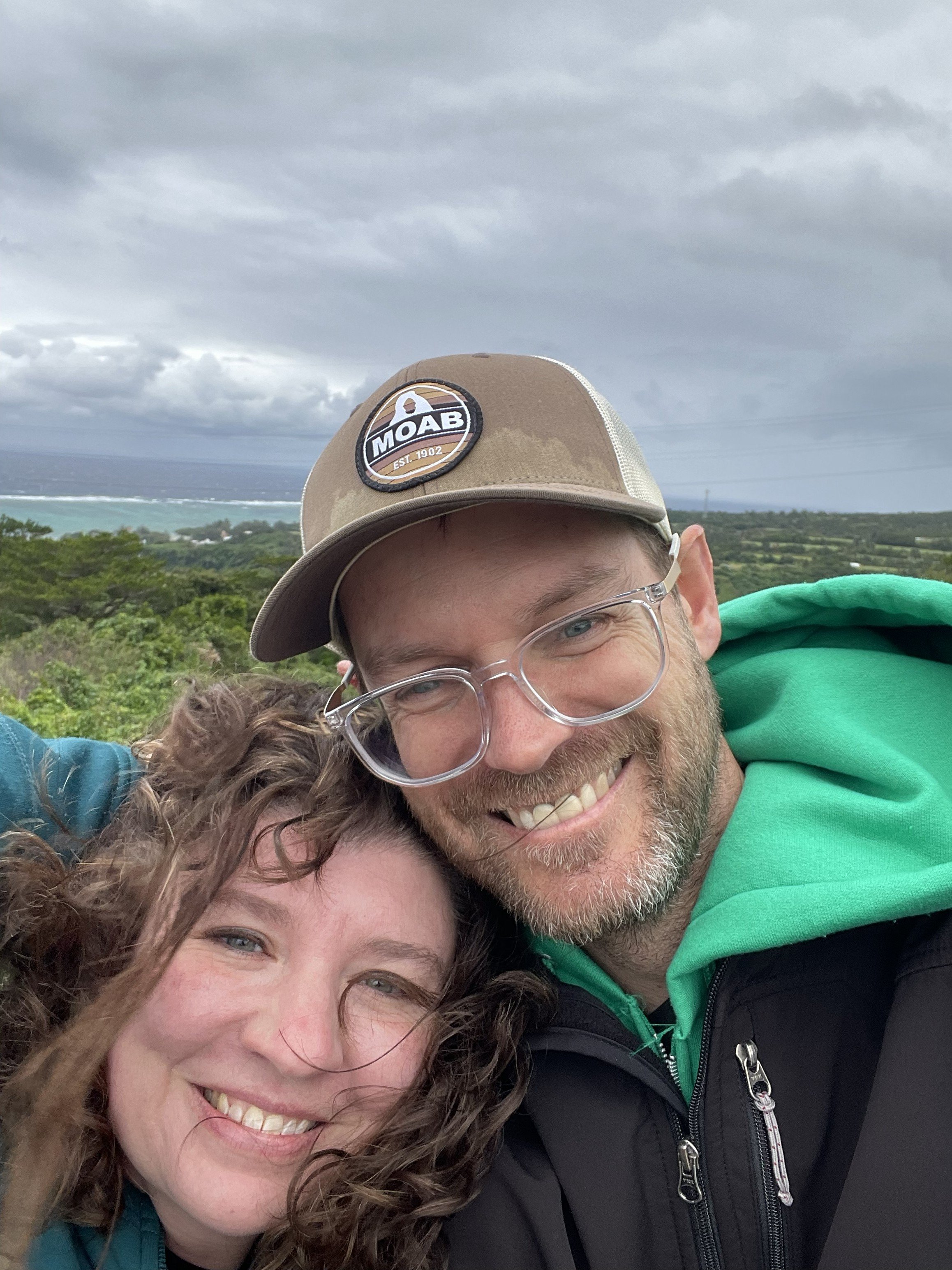 Close-up of a smiling man and woman embracing outdoors with cloudy sky and landscape in the background.
