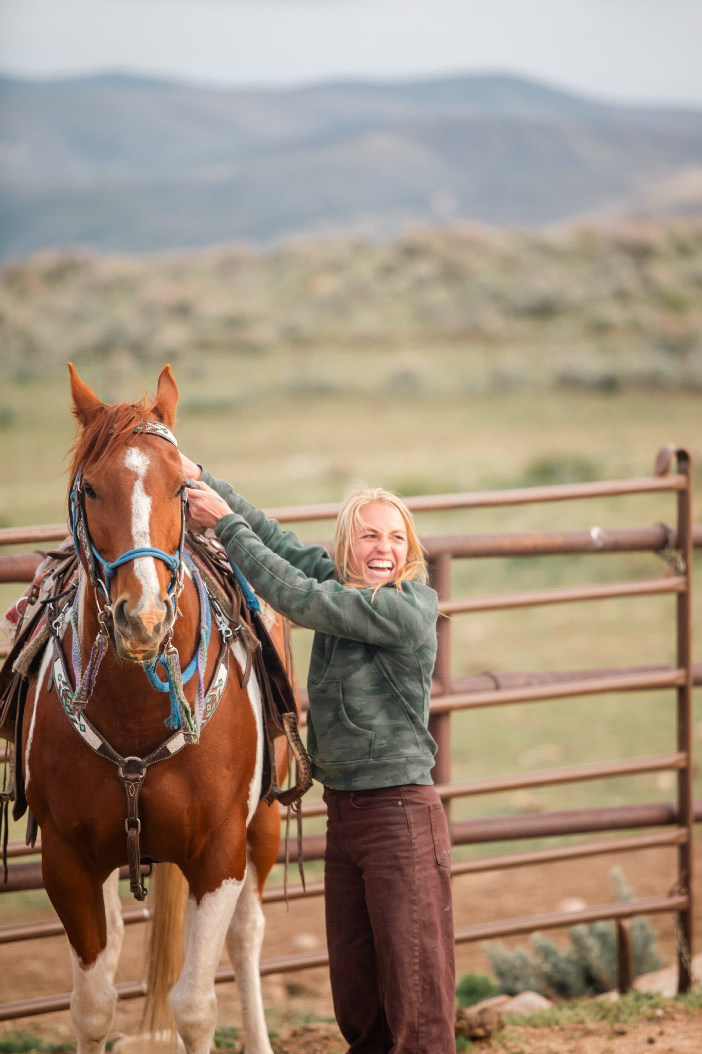 A woman with blonde hair in a green camouflage jacket and brown pants standing next to a chestnut horse with white markings on its face, in an outdoor riding arena with a wooden fence and mountainous terrain in the background.