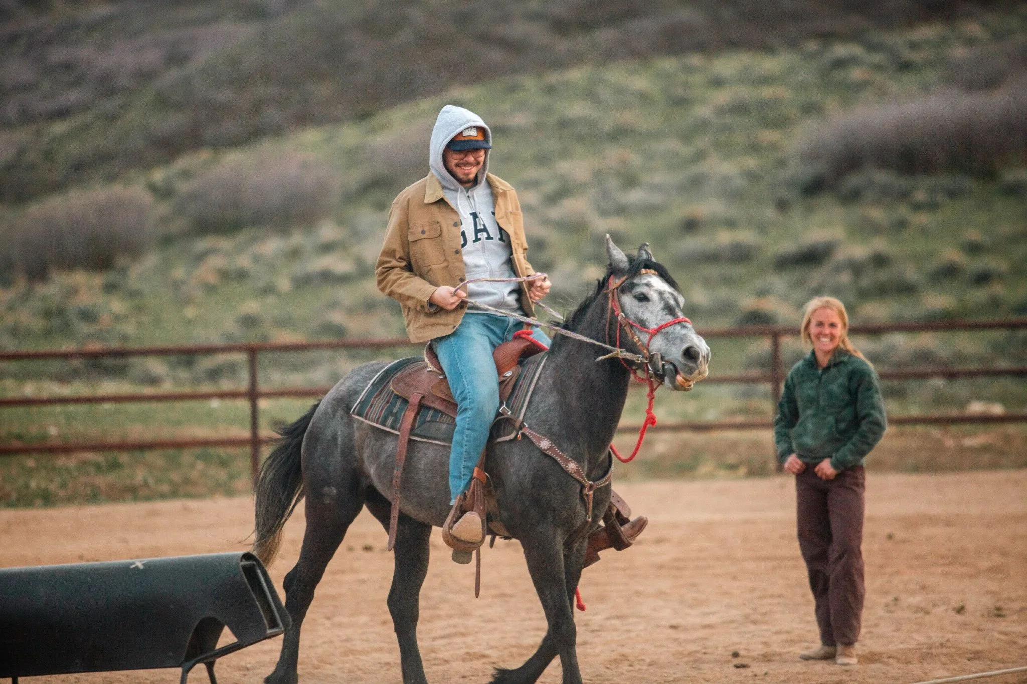 A man riding a horse in an outdoor arena, smiling, wearing a gray hoodie, tan jacket, and sunglasses, with a woman giving him a horseback riding lesson, with a blurred natural background.