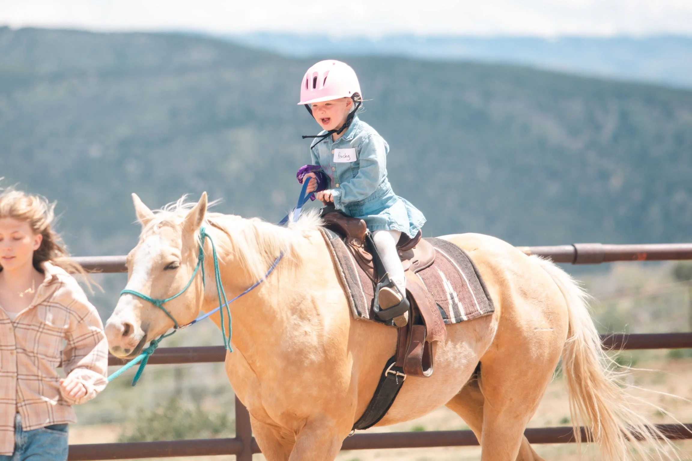 Young girl wearing a pink helmet riding a light-colored horse, held by a woman in plaid shirt, in a riding arena with mountains in the background during a kid's horseback riding lesson.