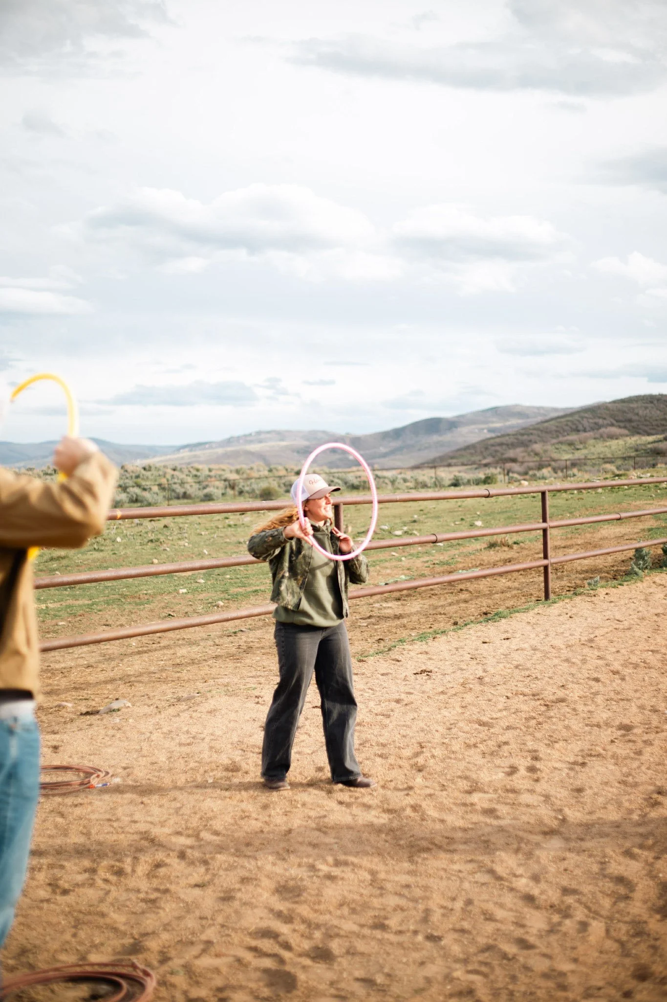 A woman standing outdoors holding a pink hula hoop, teaching people how to rope, with mountains and cloudy sky in the background.
