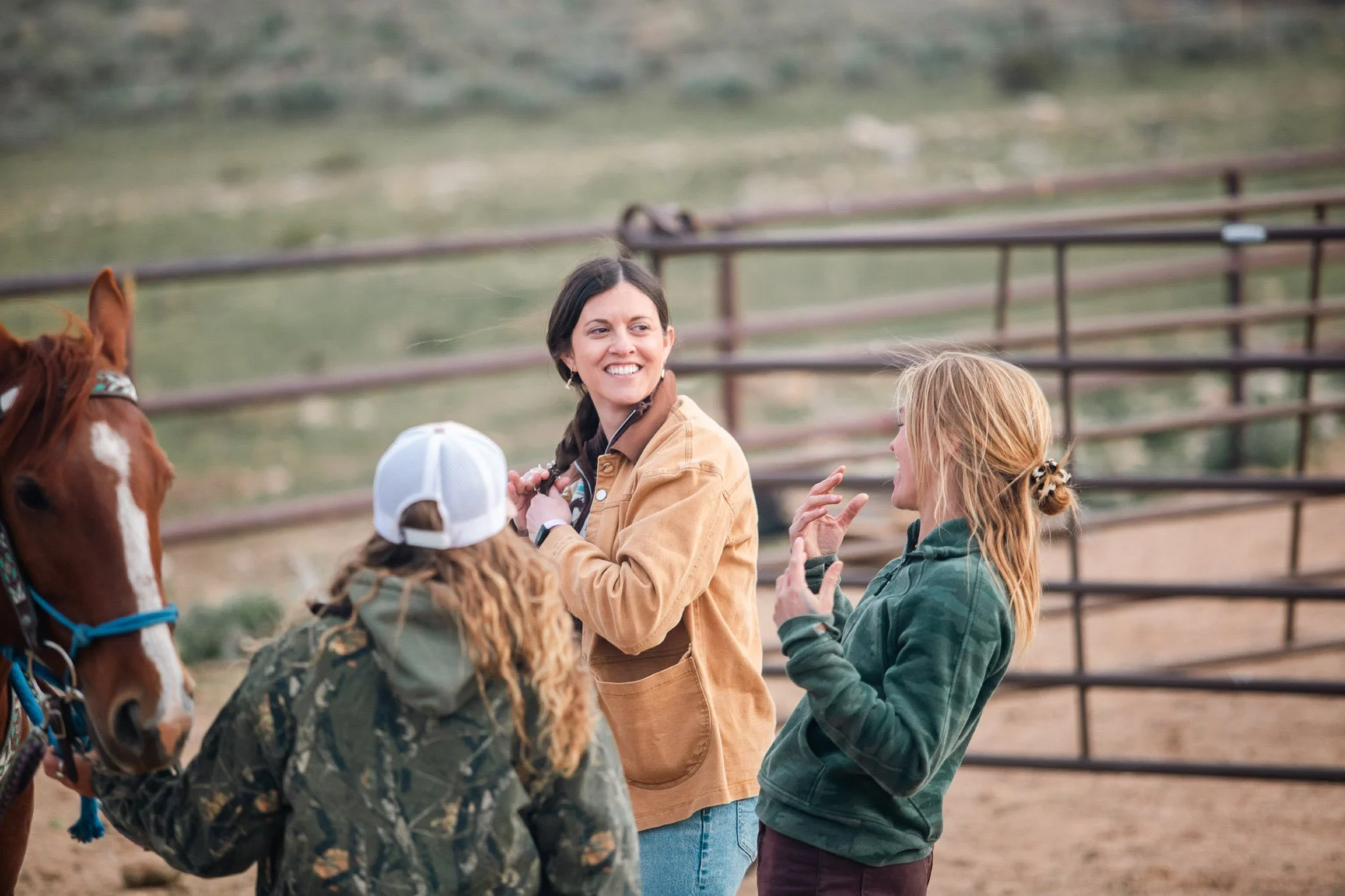 A group of children and a woman interacting with a brown and white horse at a ranch or farm, with a wooden fence and hills in the background during a ranch experience.