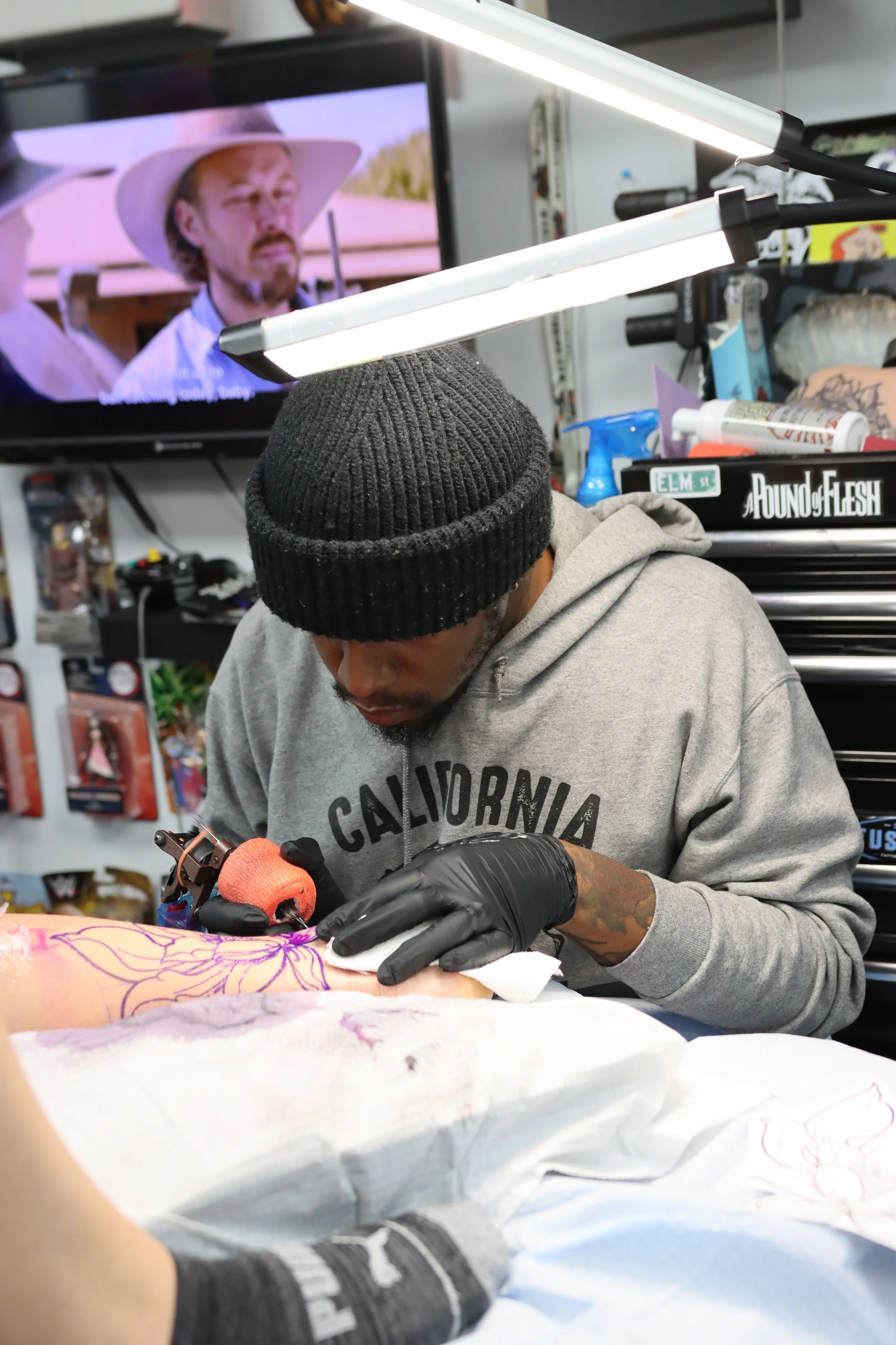 A tattoo artist with a black beanie, gray hoodie with the words 'SAN FRANCISCO' on it, and black gloves, working on a design on a client's arm in a tattoo shop.