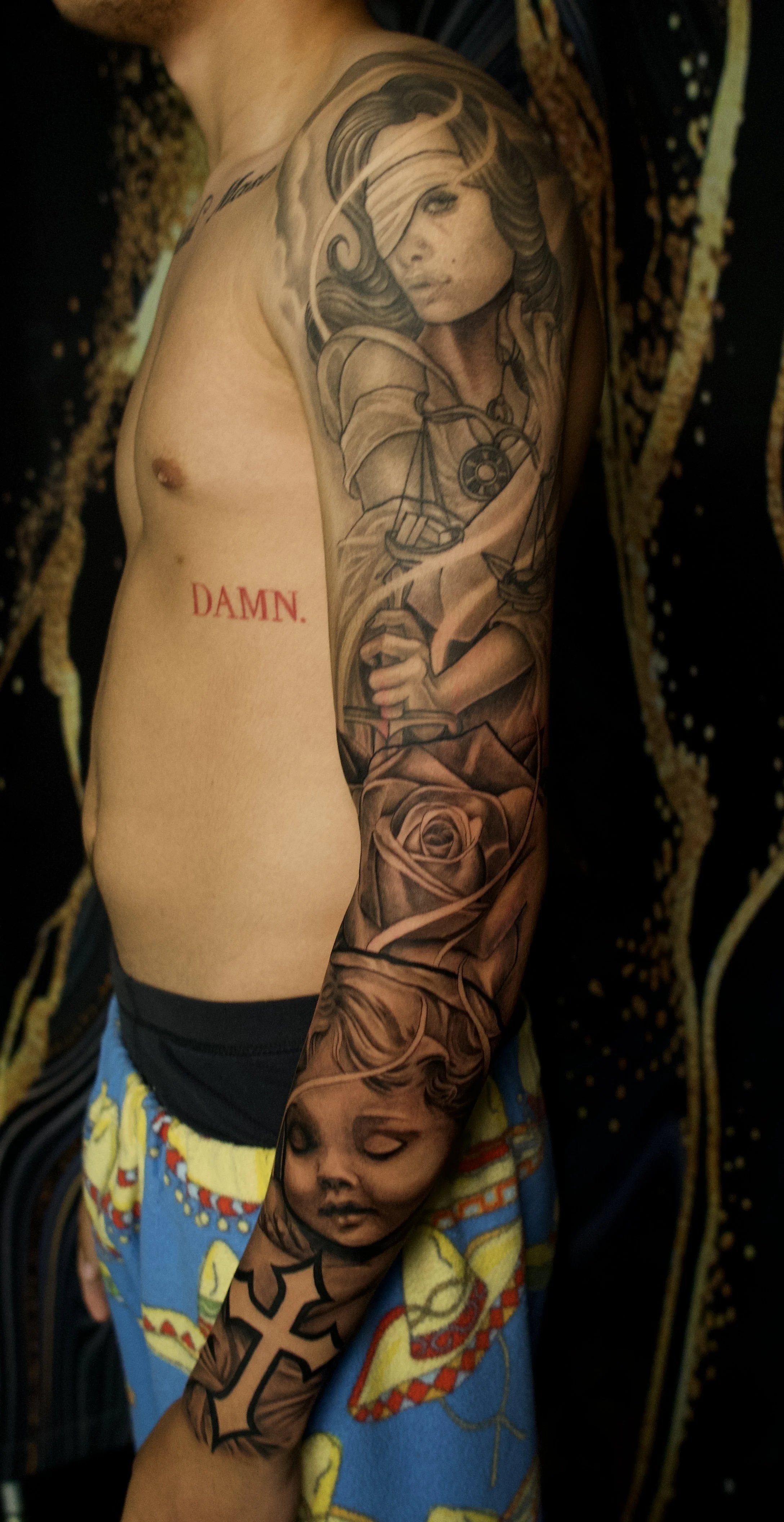 A man's arm with a large tattoo of a woman with flowing hair, holding scales of justice, and a rose at the bottom. The tattoo includes shading and detailed line work. The man is wearing colorful shorts with a cartoon pattern and has the word 'DAMN.' in red ink on his side.