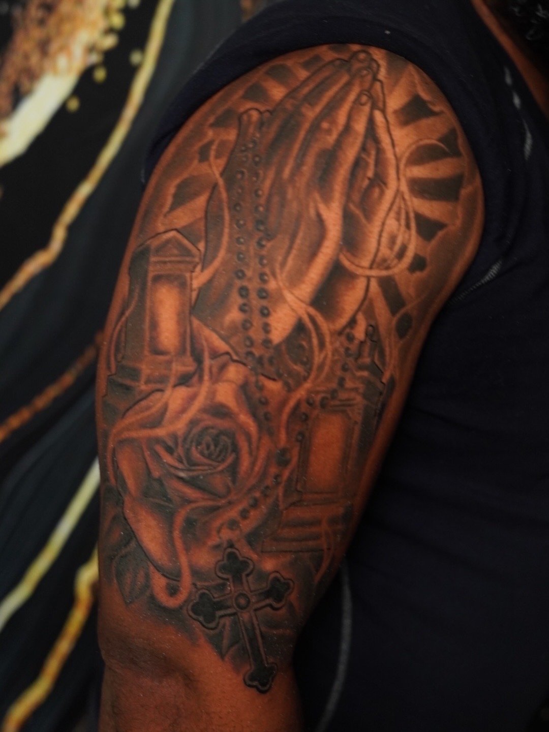 Tattoo of praying hands, a rosary, and a cross on a person's upper arm.