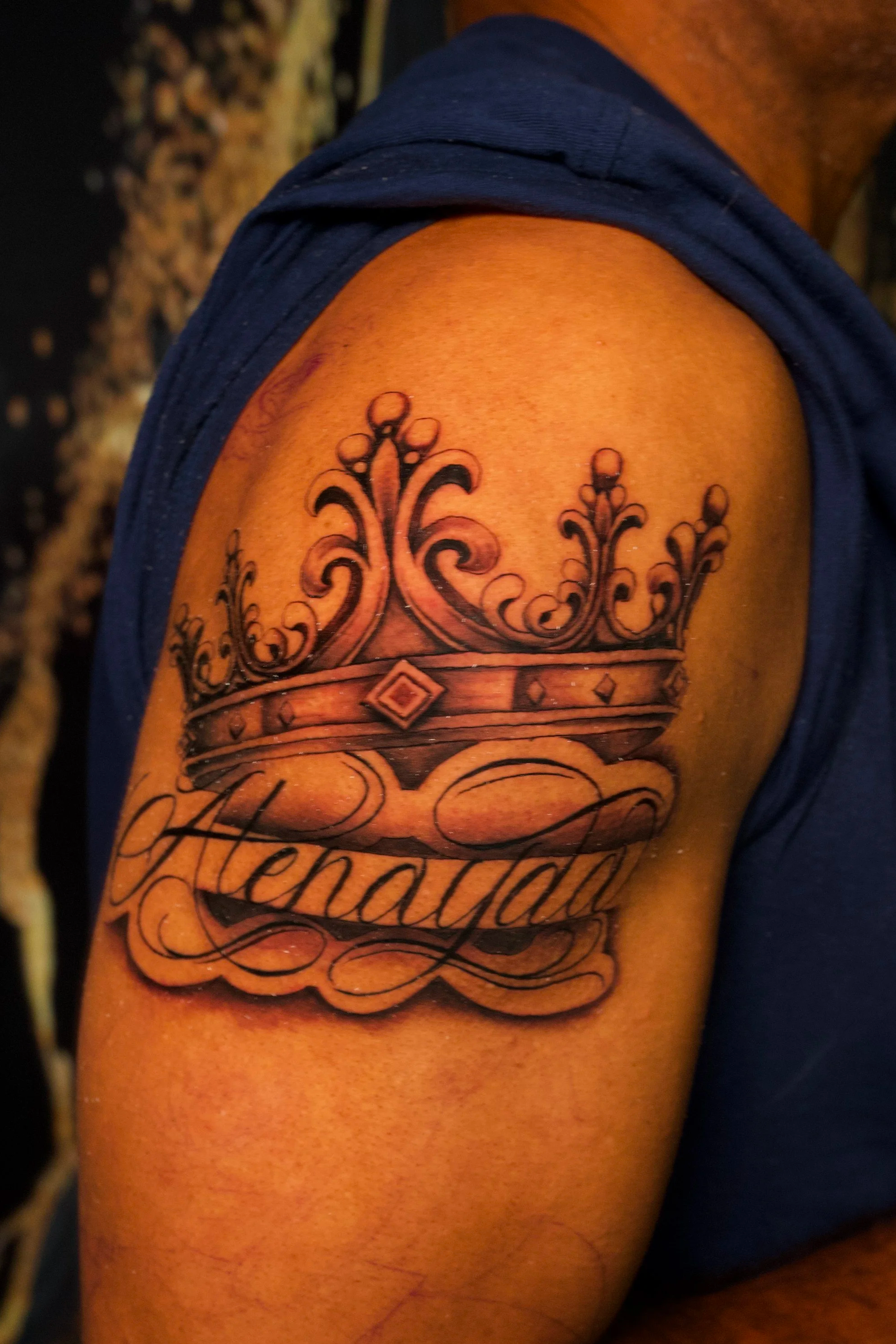 Close-up of a person's upper arm with a black tattoo featuring a crown and decorative elements, and partially visible text below it. The person is wearing a dark blue sleeveless shirt.