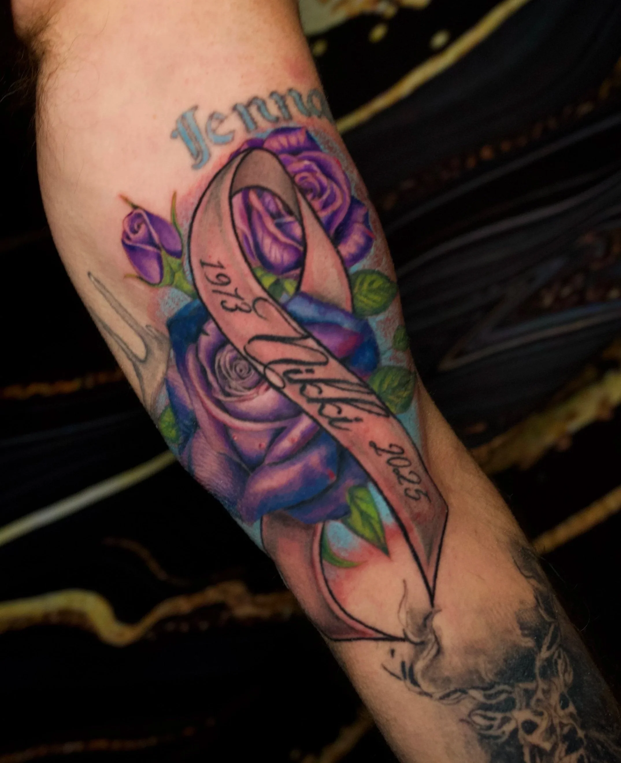 Tattoo on the forearm featuring purple roses, a pink ribbon with the year '1977', and the word 'Mom' written above.