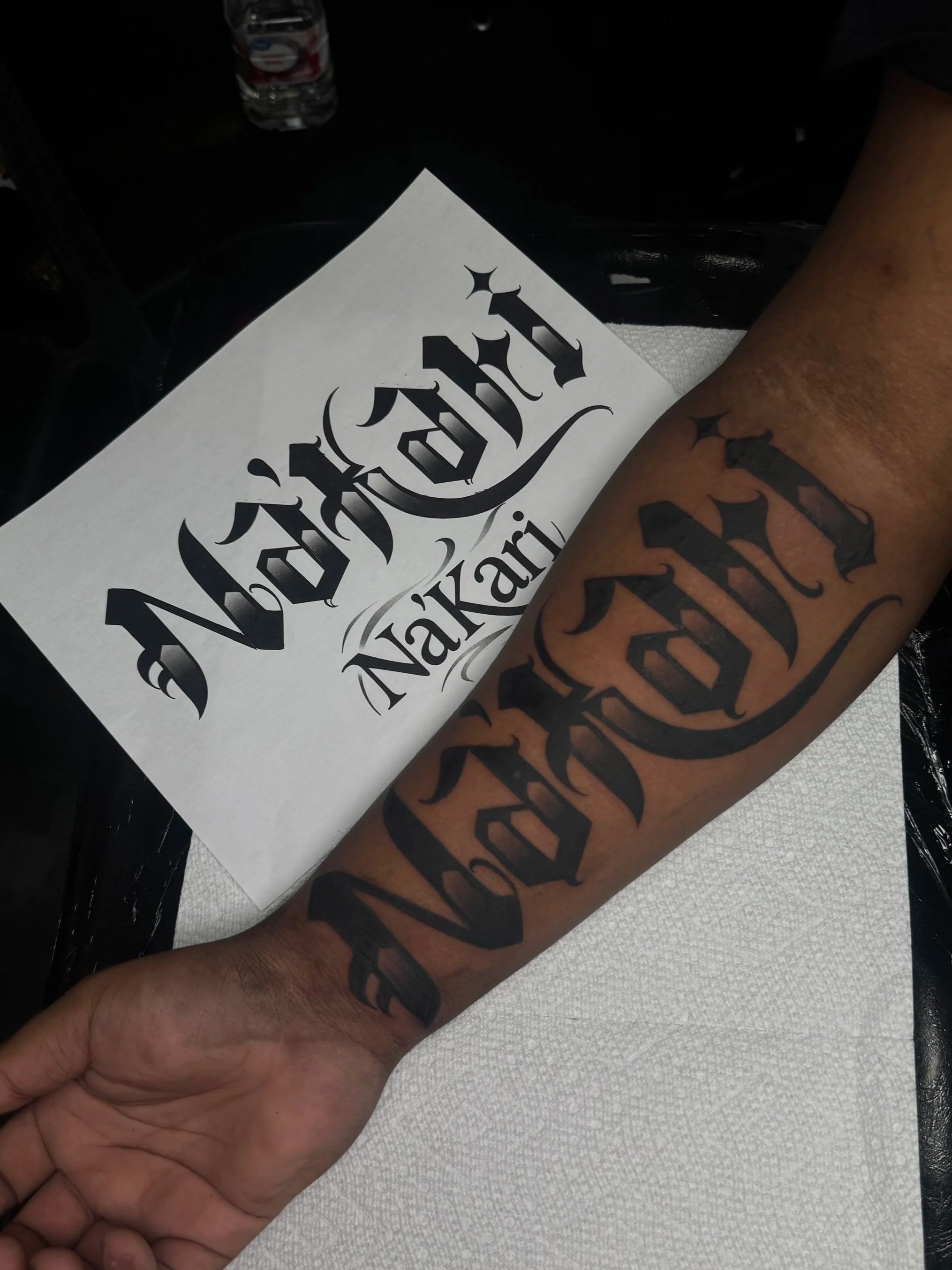 A person with tattoos on their arm, holding a paper with the word 'Nashville' in a tattoo-style font. There is a water bottle in the background.