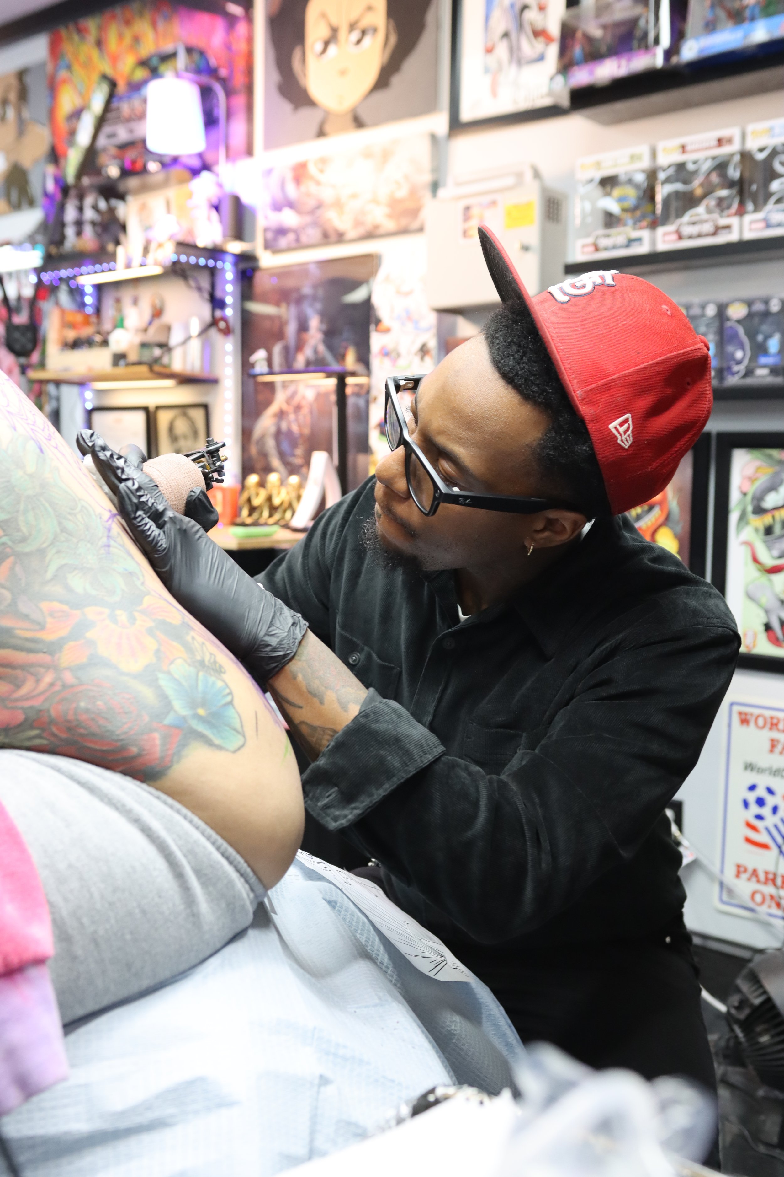 A tattoo artist working on a person's arm in a tattoo shop, surrounded by colorful artwork and tattoo supplies.
