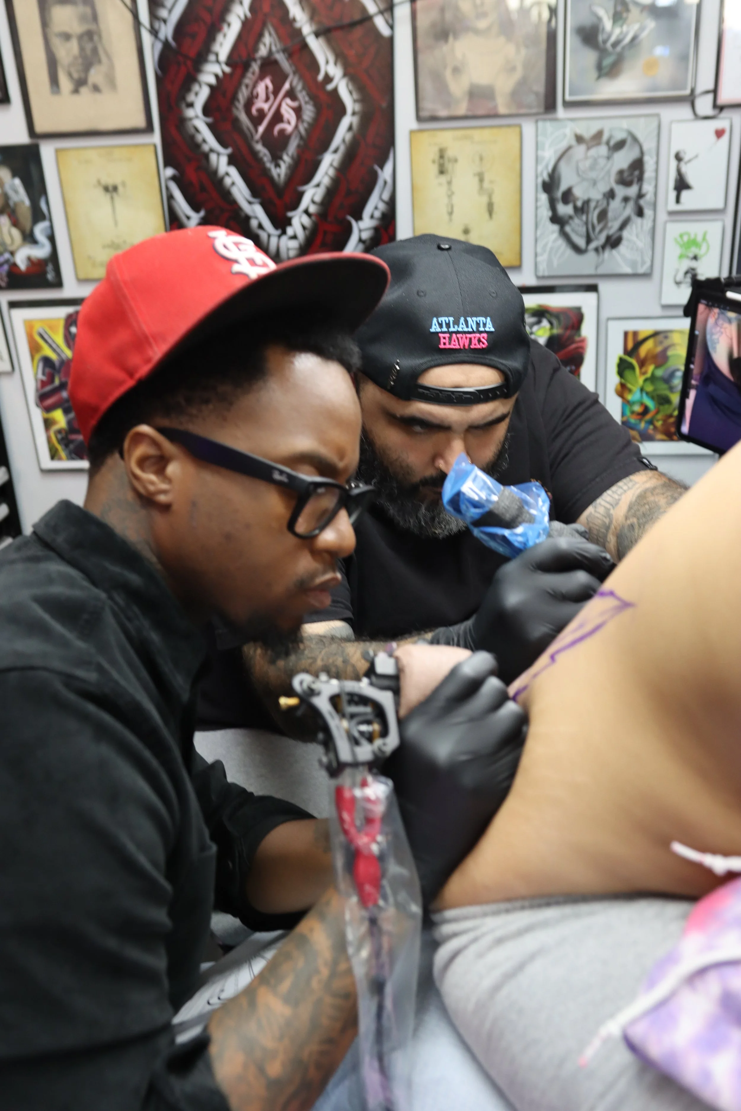 Two men with tattoos and tattoos on their arms tattooing a person. The person receiving the tattoo is not fully visible. The background features various framed art pieces on the wall.